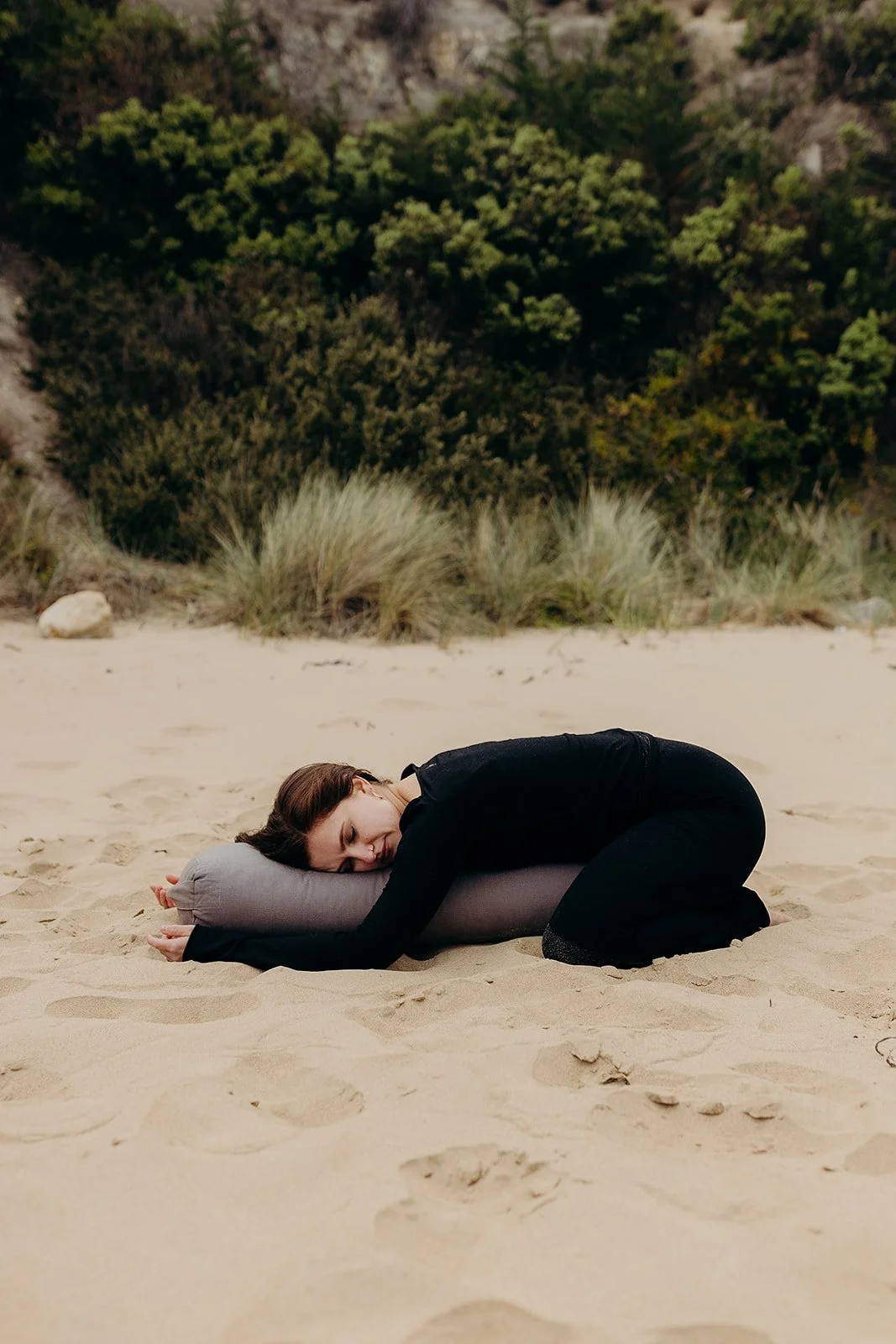 A woman in black clothing resting in child's pose on a gray bolster pillow on a sandy beach in Tasmania, hugging the bolster pillow with her eyes closed and a gentle smile, with sandy dunes and trees in the background.