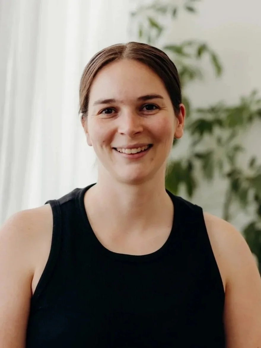 A woman with brown hair, wearing a black sleeveless top and a silver necklace with a pearl, smiling at the camera, with green plants and light-colored curtains in the background, at Willow Massage and Wellness in Howrah, Tasmania.