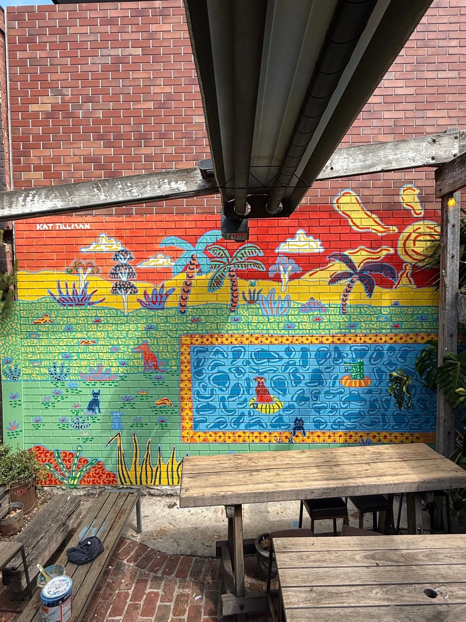 Colourful outdoor brick mural by Kat Tillman featuring palm trees, Australian plants, animals and a playful swimming pool scene.
