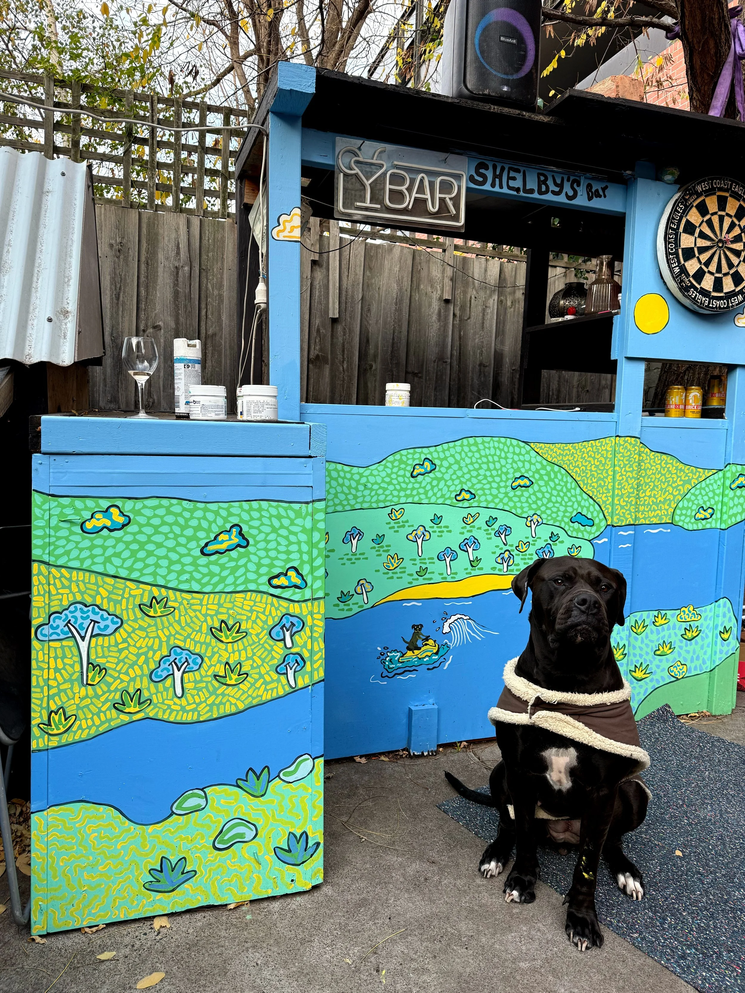 A dog sitting in front of a colorful painted outdoor bar with artwork of a landscape including hills, trees, and a river, with a dartboard, sign reading 'Shelby's Bar,' and various bottles and glasses on the bar.