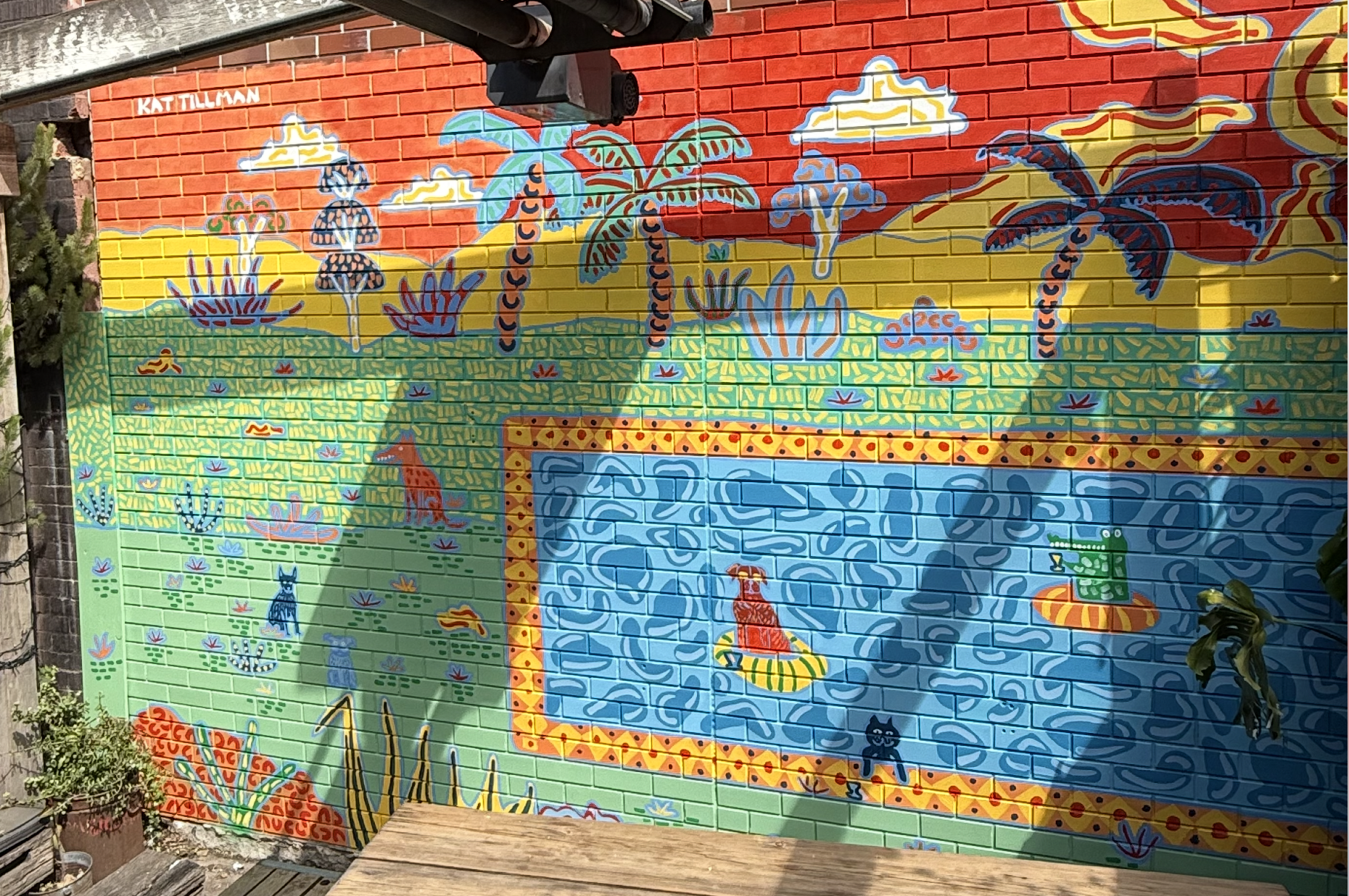 Colorful mural on a brick wall depicting a tropical scene with trees, plants, animals, clouds, and a pool. Signed by Kat Tillman.