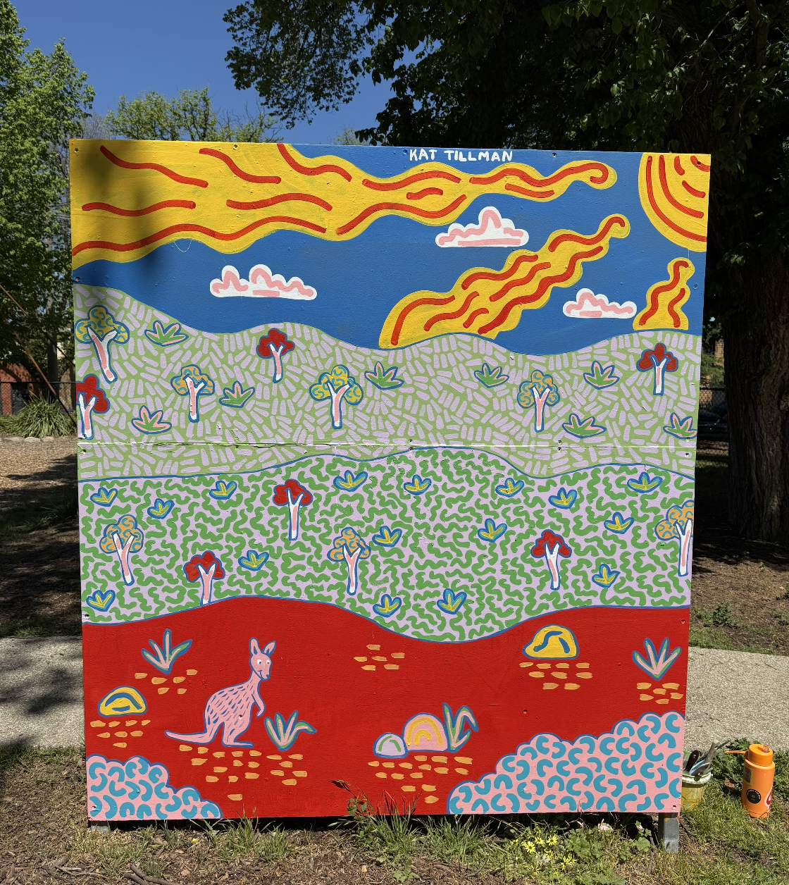 Colorful outdoor mural showing layers of landscape with stylized mountains, trees, fields, and a pink animal, likely a kangaroo, at the bottom, with the artist's name Kat Tillman written at the top.