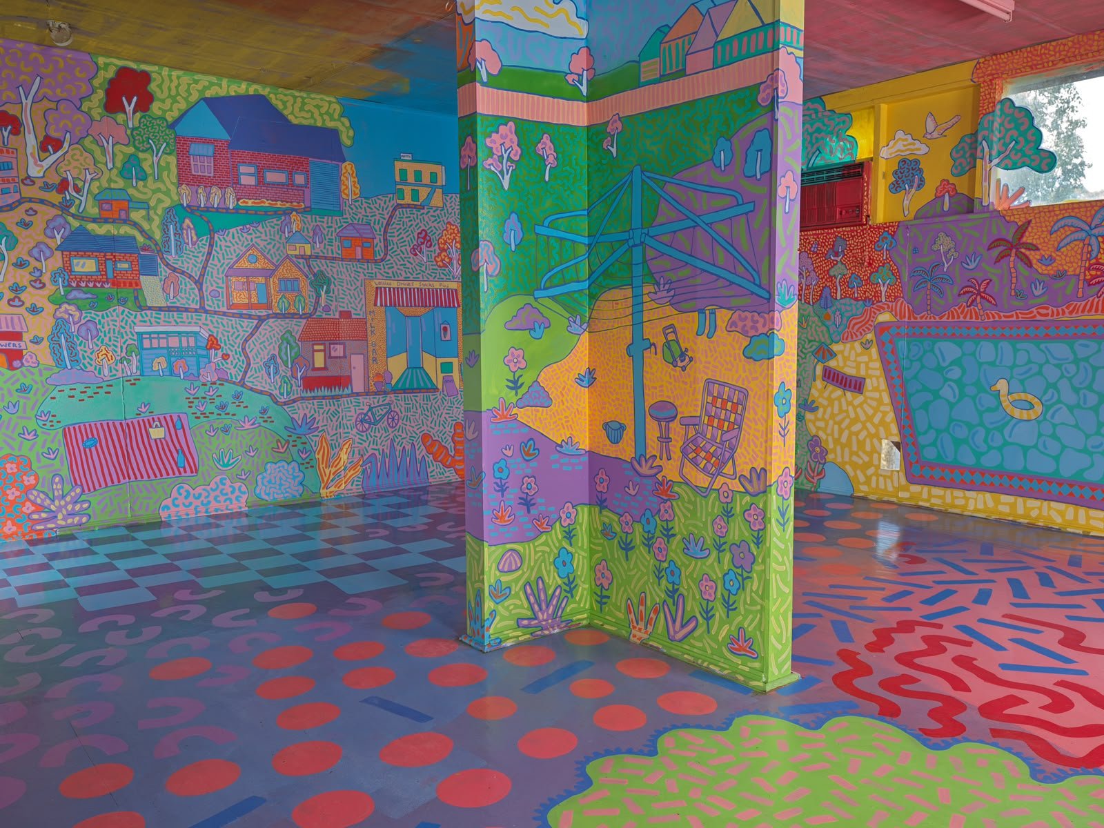 Floor-to-ceiling mural depicting colourful houses, green landscapes and a bridge, surrounded by bold geometric floor patterns inside the Side Quest Stories installation.