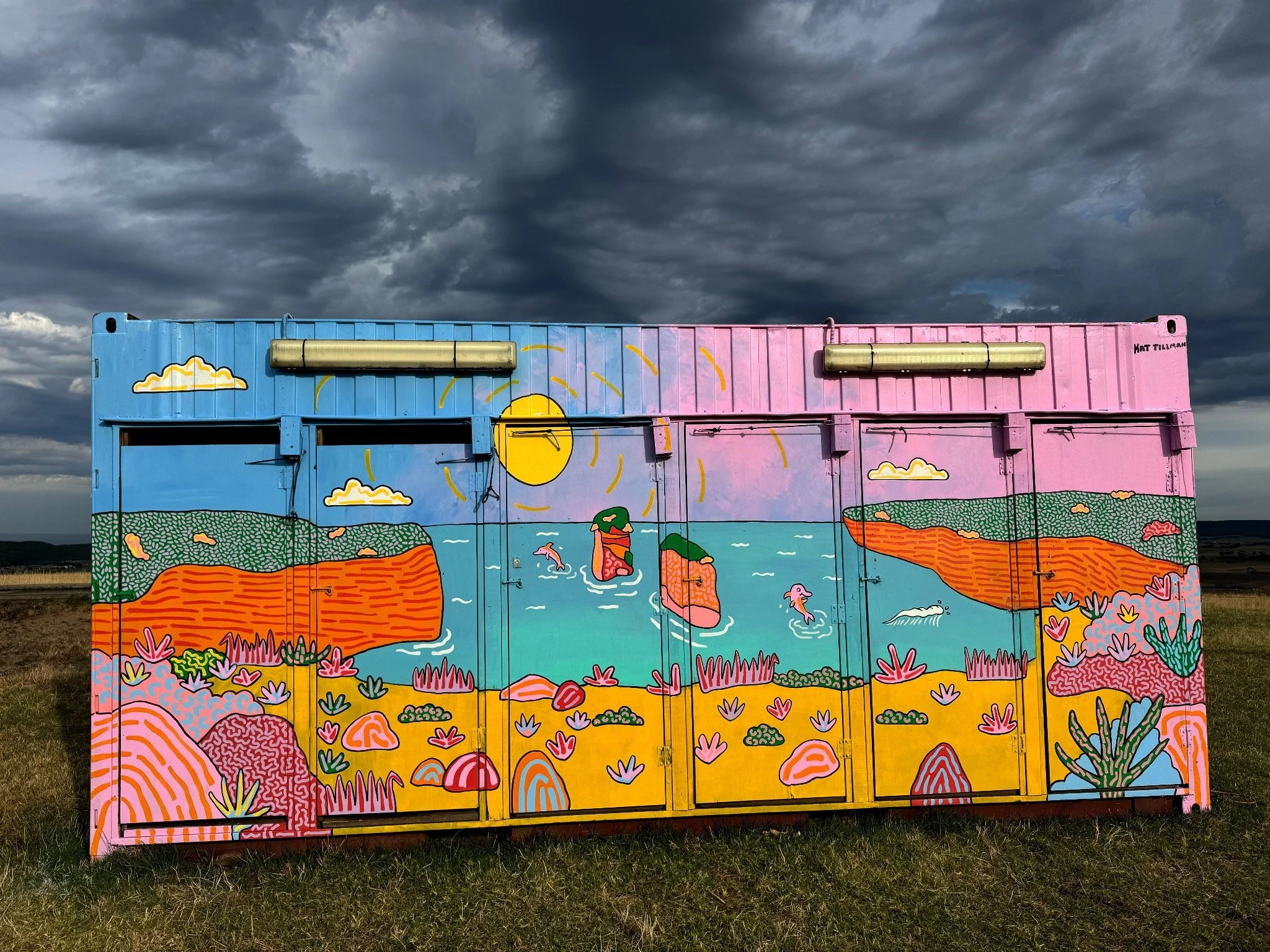 Colourful festival mural by Kat Tillman depicting an Australian coastal landscape with water, cliffs, swimmers, native plants and a setting sun.