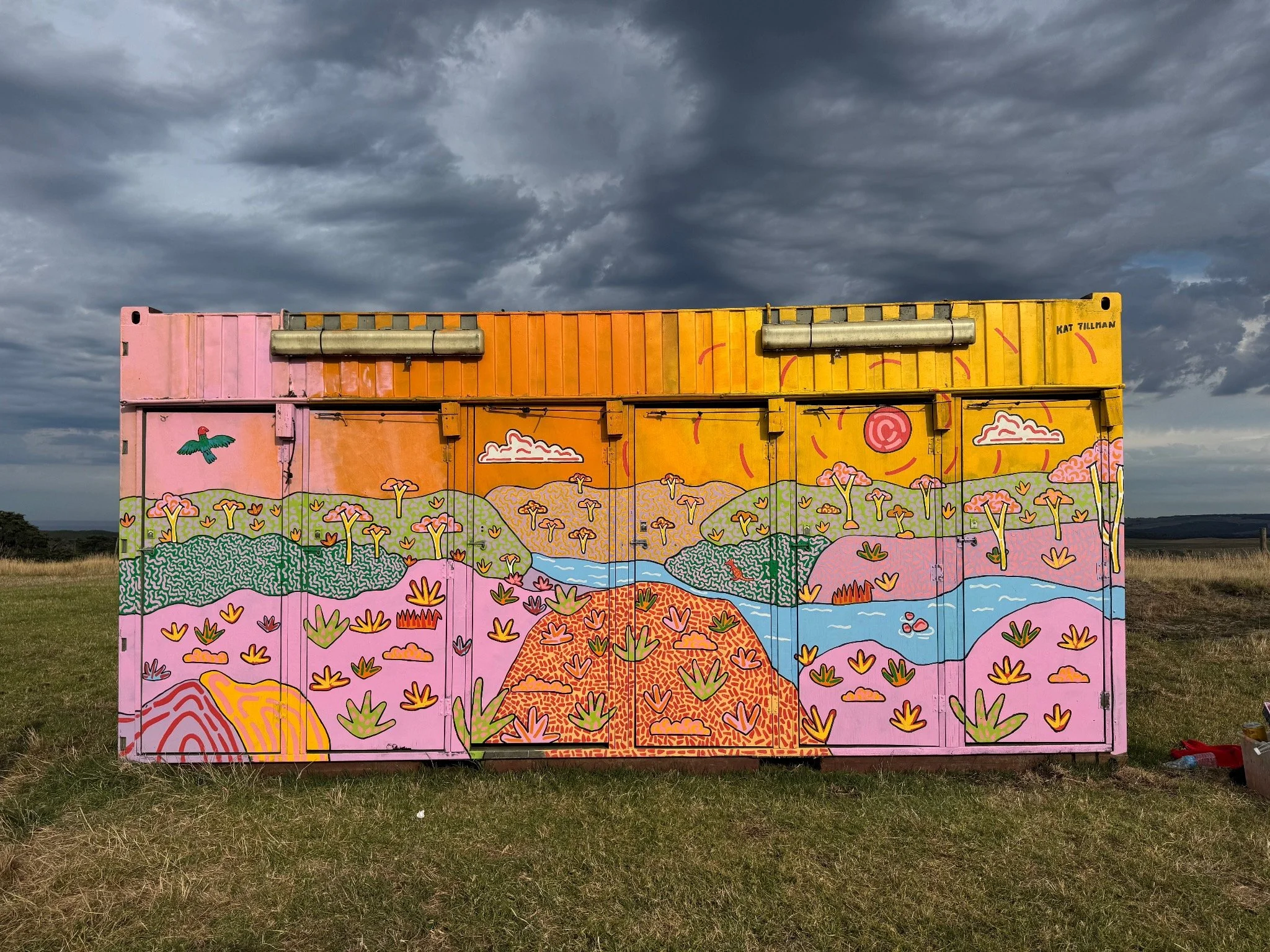 Large-scale painted container mural by Kat Tillman featuring a colourful Australian landscape beneath a dramatic cloudy sky.