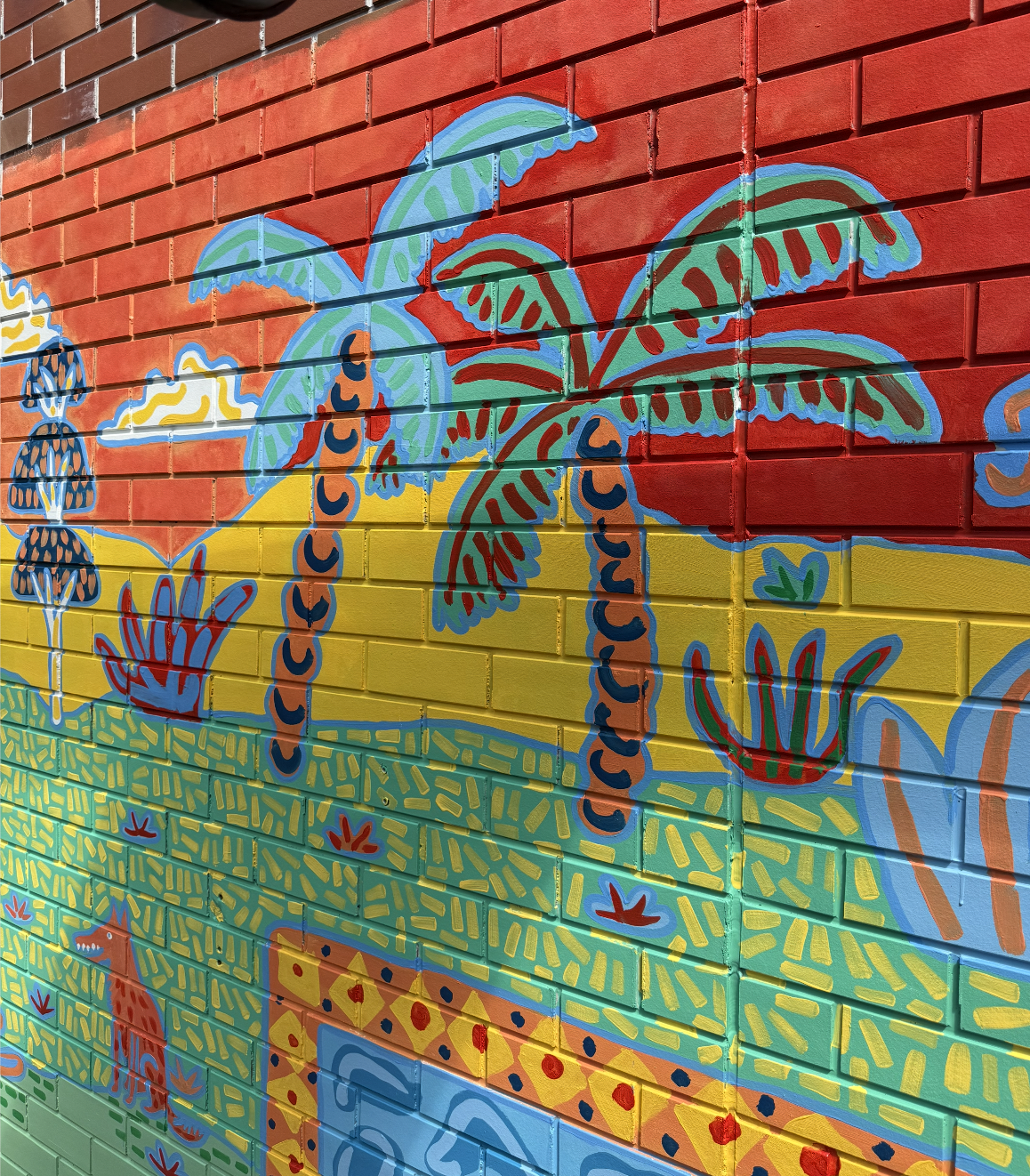Colorful mural on a brick wall depicting a tropical scene with palm trees, a sun, a dinosaur, and abstract patterns in vibrant colors.