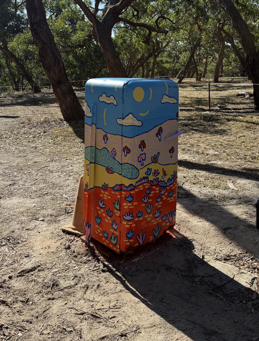 A colorful mailbox painted with a landscape scene featuring a yellow field, blue and purple flowers, green hills, and a blue sky with a yellow sun, situated among trees.