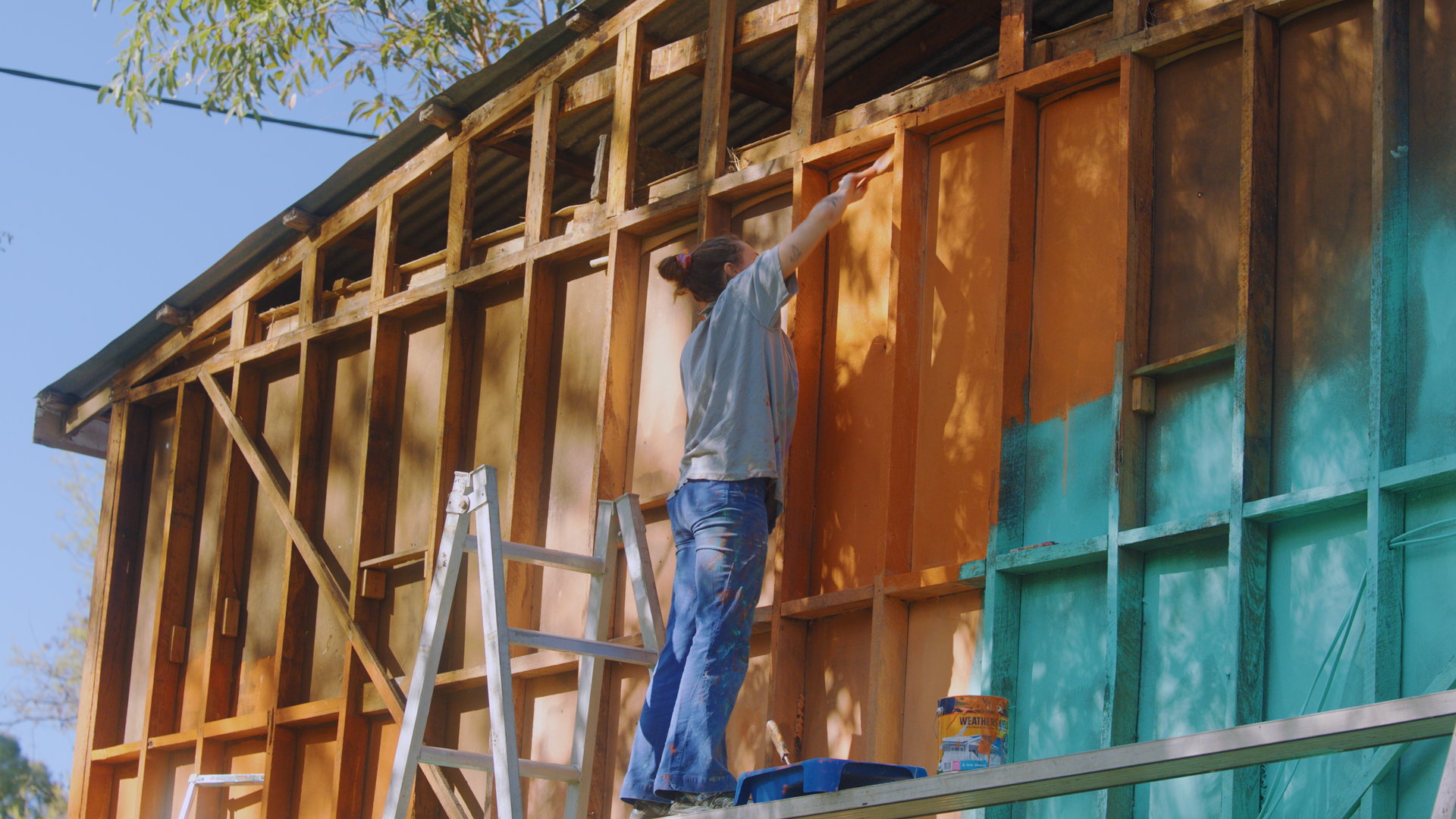 A woman standing on a ladder working on the wooden framework of a house under construction, with colorful insulation panels inside the walls.