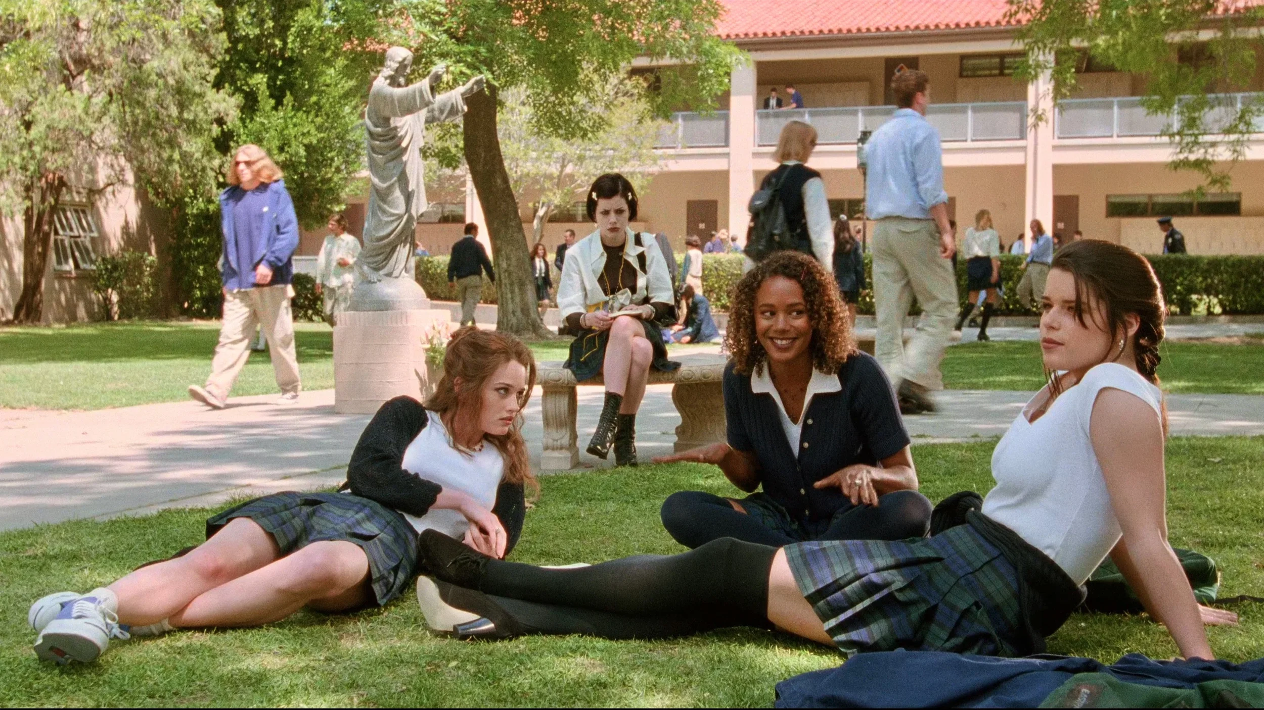 Robin Tunney, Fairuza Balk, Neve Campbell and Rachel True gather on the lawn of their Catholic high school in The Craft (1996), with other students and a religious statue in the background.