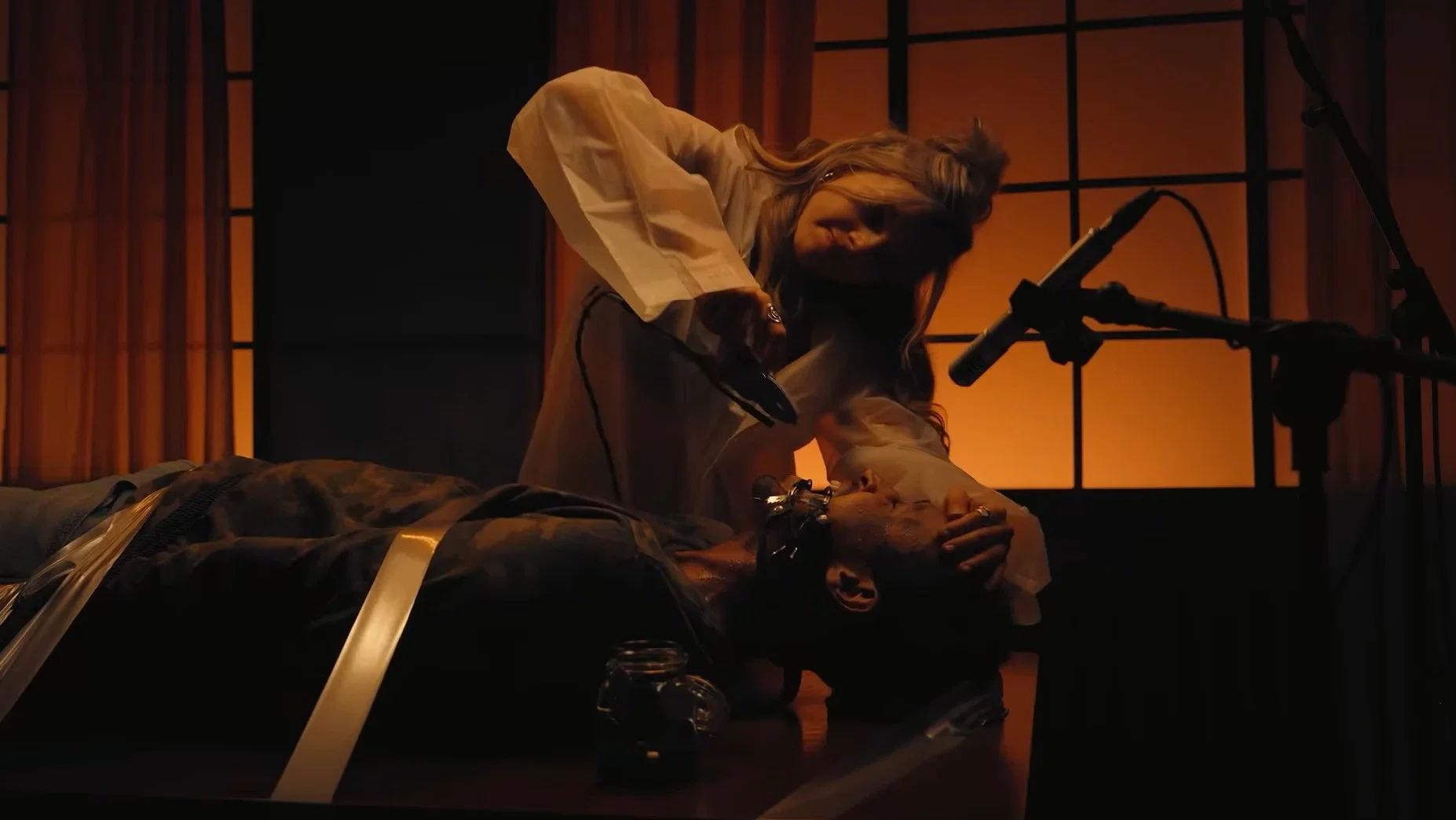 A woman leans over a man lying on a table surrounded by recording equipment and microphones in the Death Tone segment of V/H/S/Mixtape (2026).