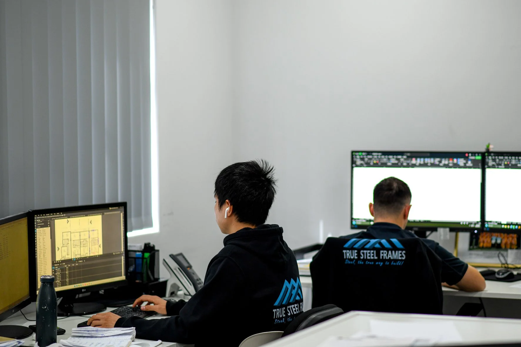Two people working on computers in an office, with multiple monitors displaying design and technical work.