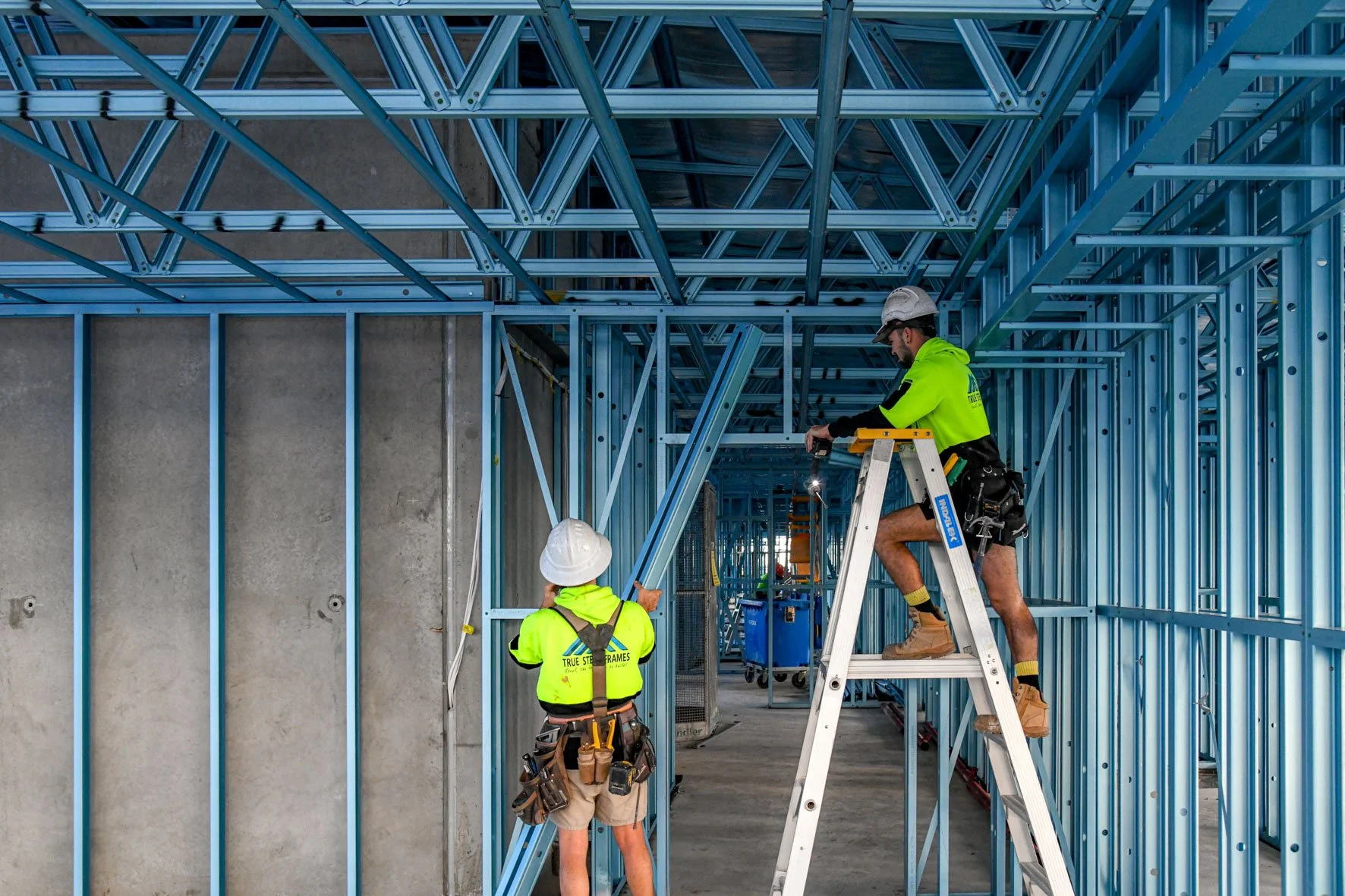 Two construction workers building a structure with blue steel framing. One worker is standing on the ground pointing, the other is on a ladder working on the framing. Both wearing high-visibility safety gear and white hard hats.