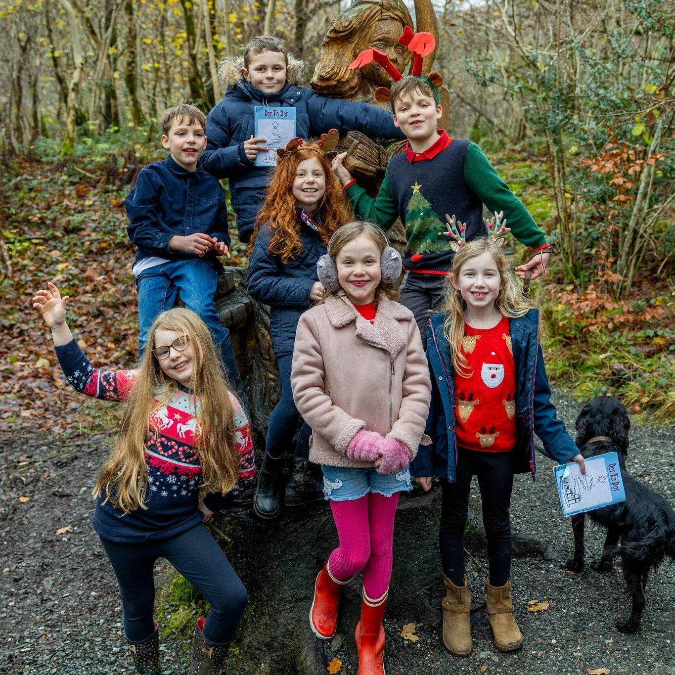Group of children and a dog at an outdoor wooded location dressed in festive and winter clothing, posing in front of a wooden sculpture of a person's face with reindeer antlers.