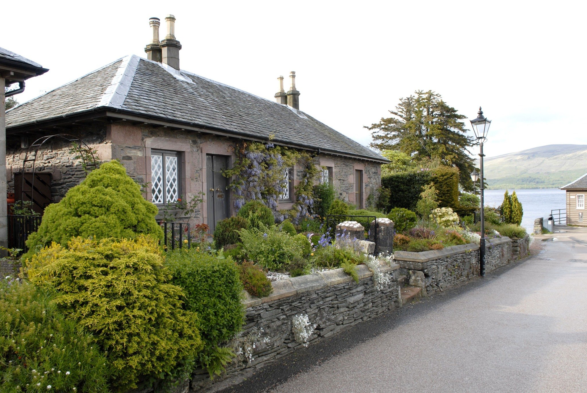 A charming stone cottage with a garden, overlooking a body of water with hills in the distance.