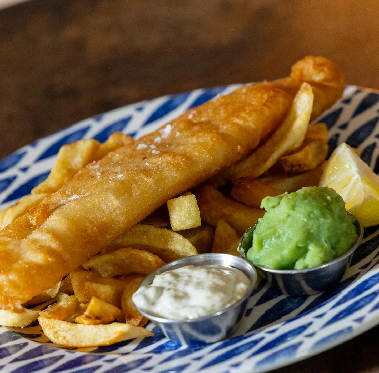 A plate of fish and chips with lemon wedges, tartar sauce, and guacamole on a patterned blue and white dish.