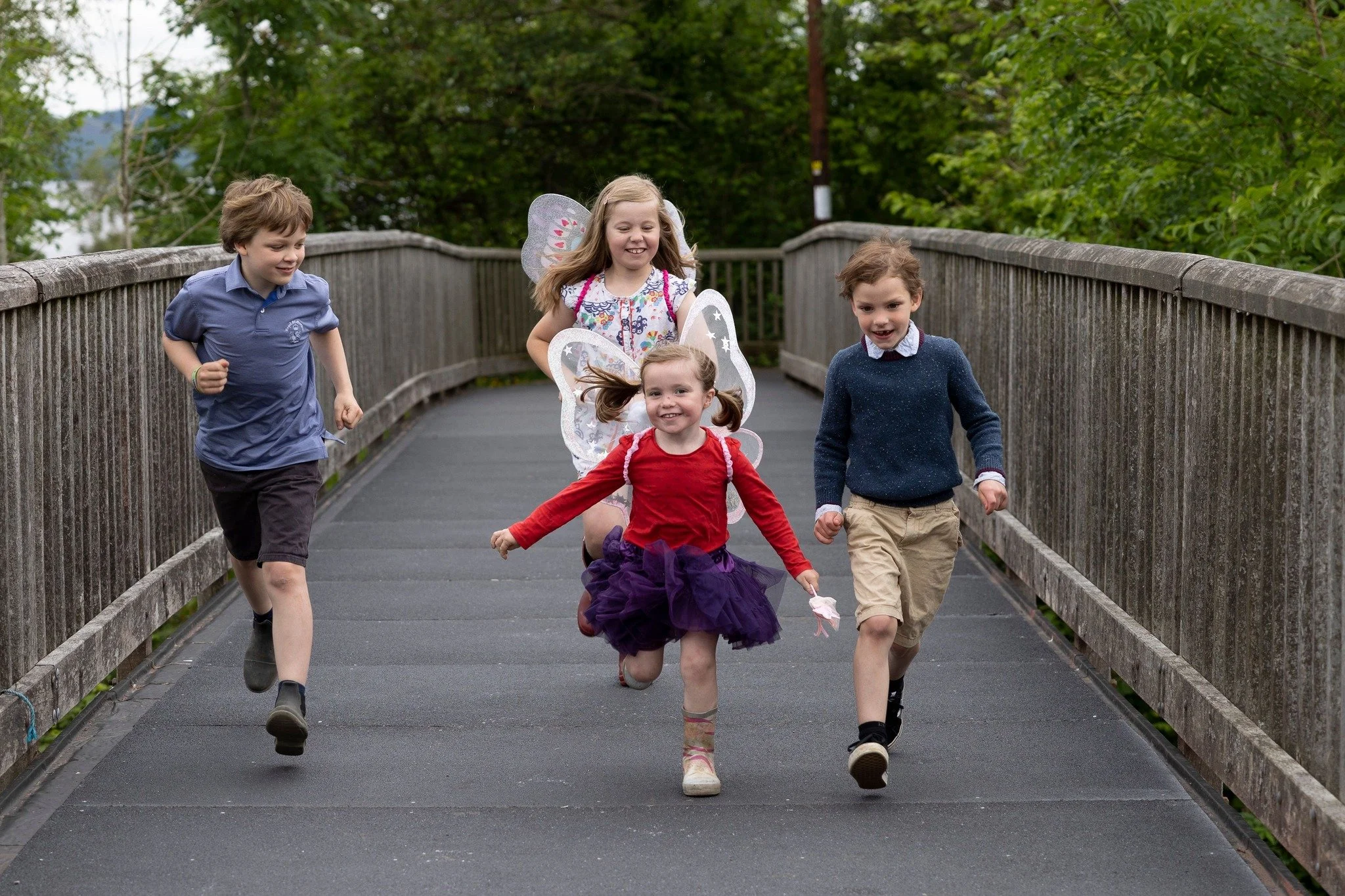 Four children running and playing on a wooden bridge, with two girls dressed as fairies with wings and a purple skirt, and two boys in casual clothes, surrounded by green trees.