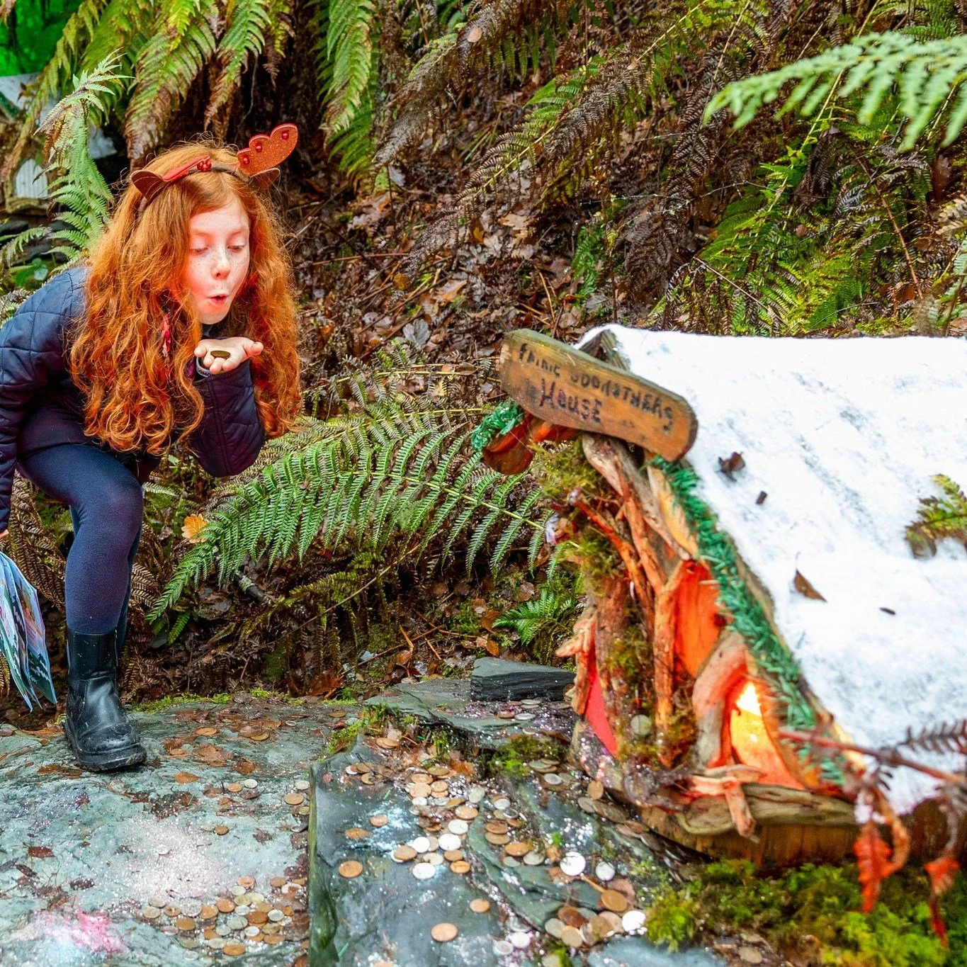 A young girl with red hair wearing reindeer antlers hat, blowing coins into a small wooden house labeled 'Little House' in an outdoor forest setting with ferns and moss.