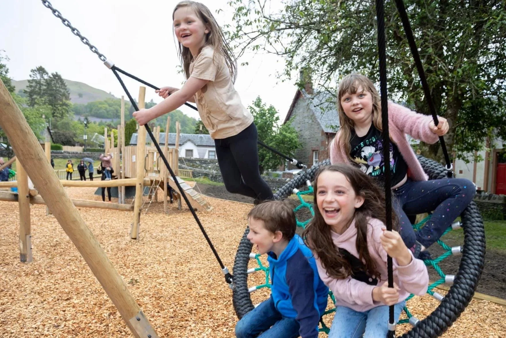 Four children playing on a large round outdoor swing at a playground, with a wooden play structure and trees in the background, smiling and enjoying the activity.