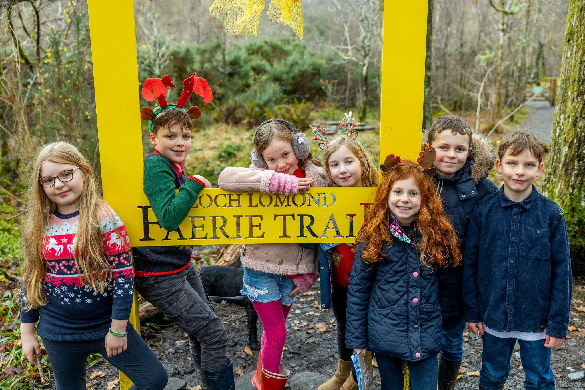 Group of children at a forest trail, wearing reindeer antlers and Christmas sweaters, people leaning on a yellow frame with a sign that reads "Loch Lomond Fairy Trail", trees and greenery in the background.