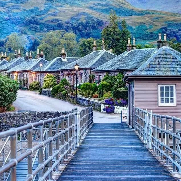 A view of a village with row of houses, a wooden footbridge in the foreground, lush greenery, flowers, and hills in the background.