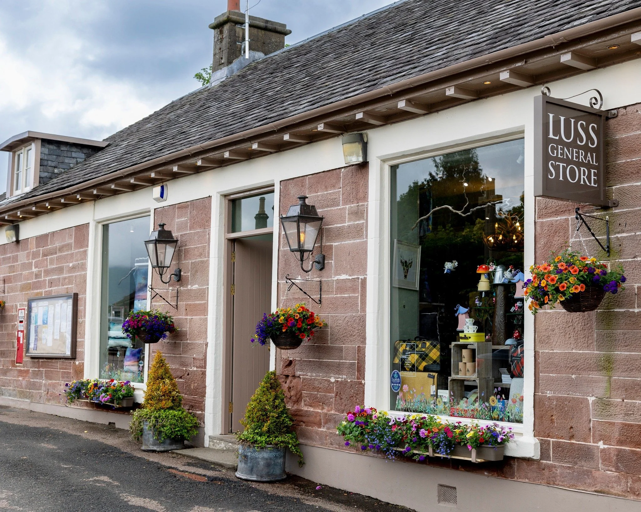 Front view of a brick store with a sign reading 'Luss General Store', decorated with hanging and potted colorful flowers, black lantern-style lights, and a bulletin board outside.