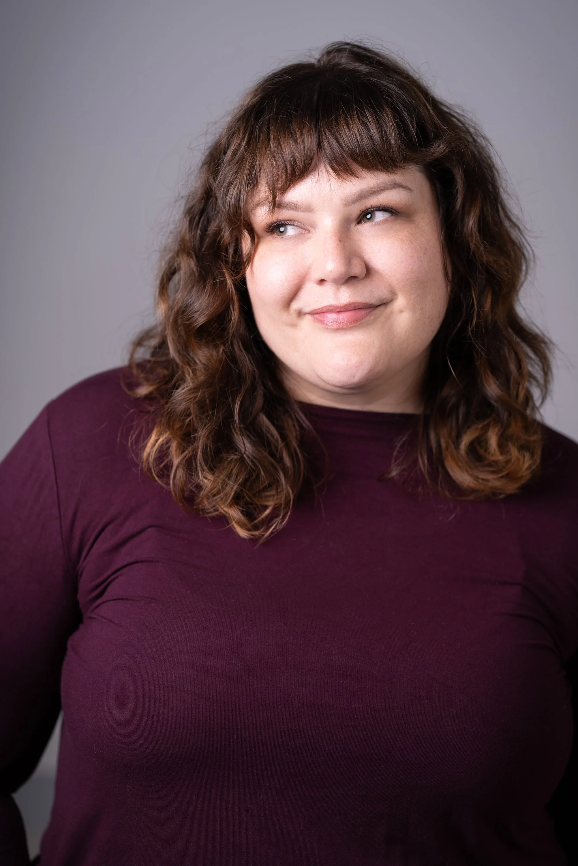 Close-up of a woman with medium length brown wavy hair, wearing a purple top, smiling softly against a neutral background.