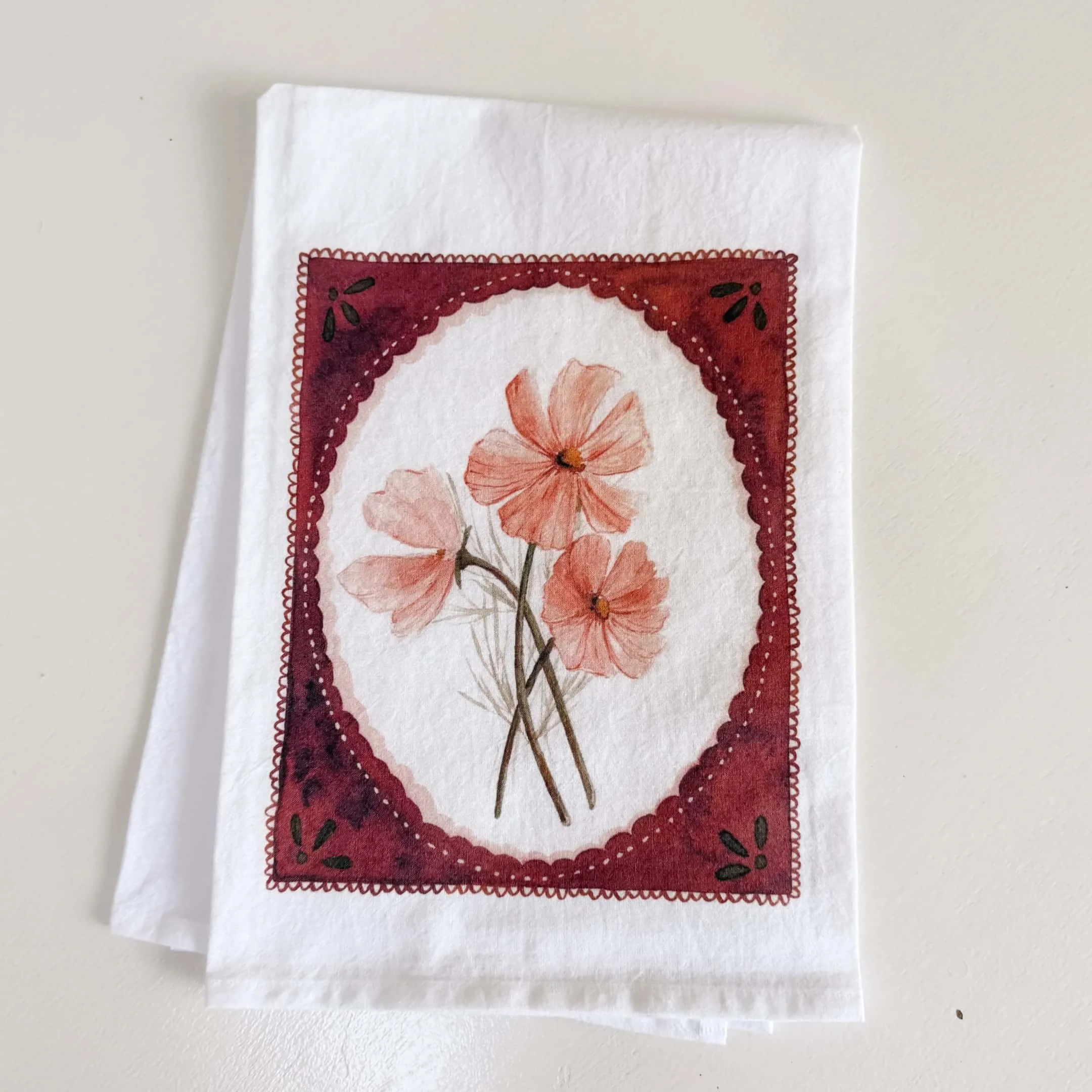 Hand-painted pink cosmos watercolor tea towel with a rust scalloped border, folded flat on a white background