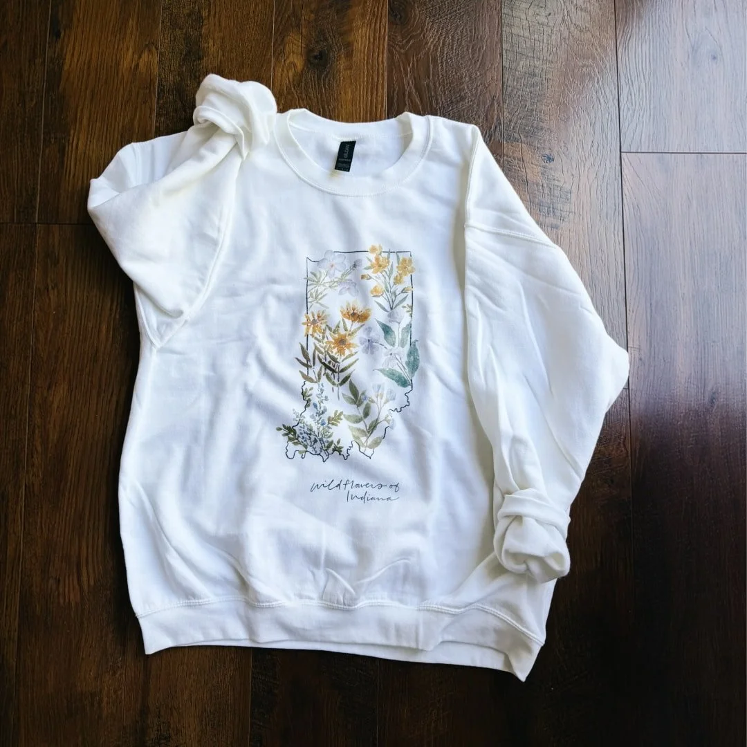 State of Indiana wildflowers watercolor sweatshirt: white crewneck with wildflowers inside the Indiana outline, laid flat on a wood floor