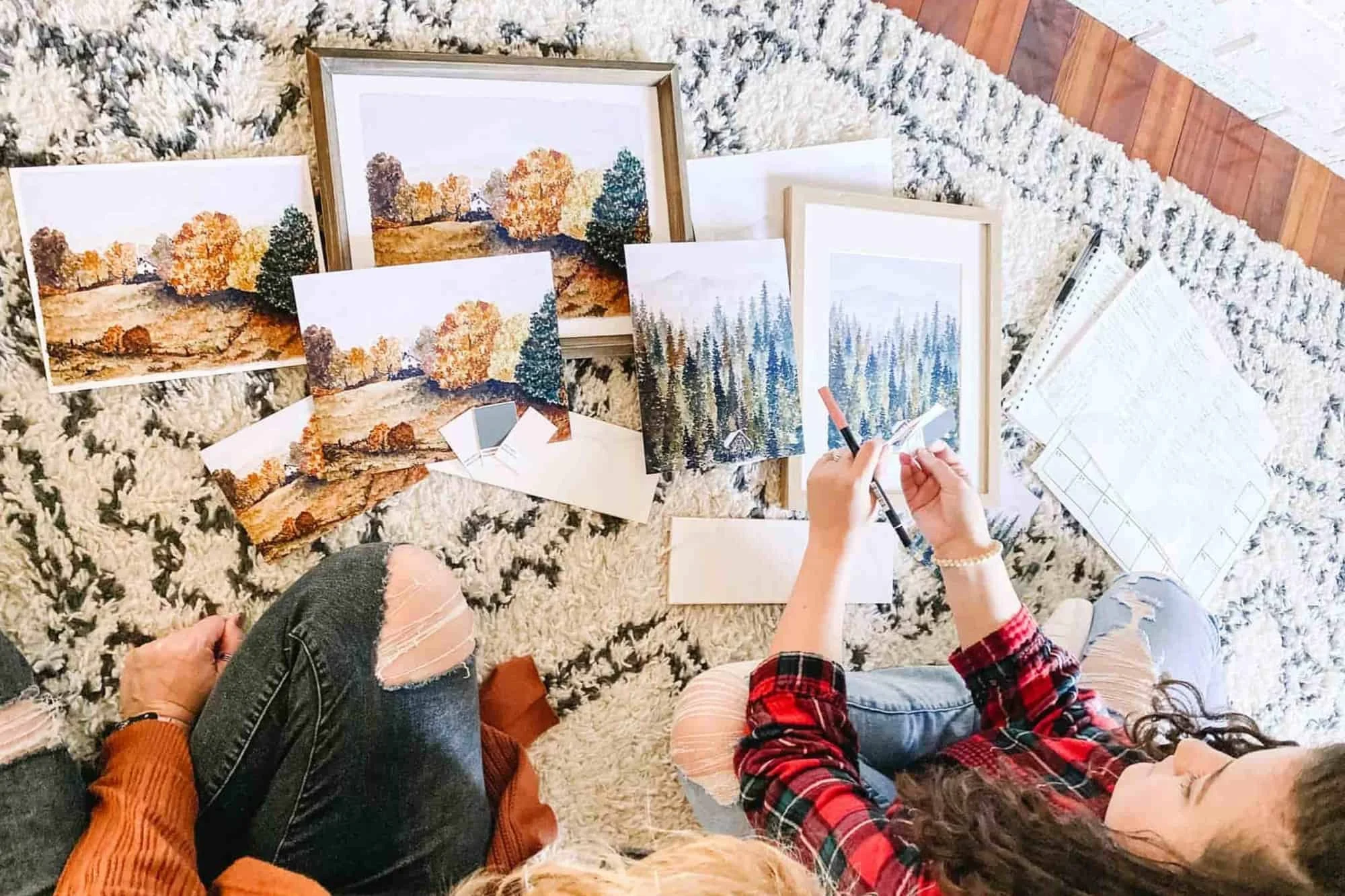 Two people sitting on a rug choosing between framed and unframed watercolor landscape art prints laid out around them