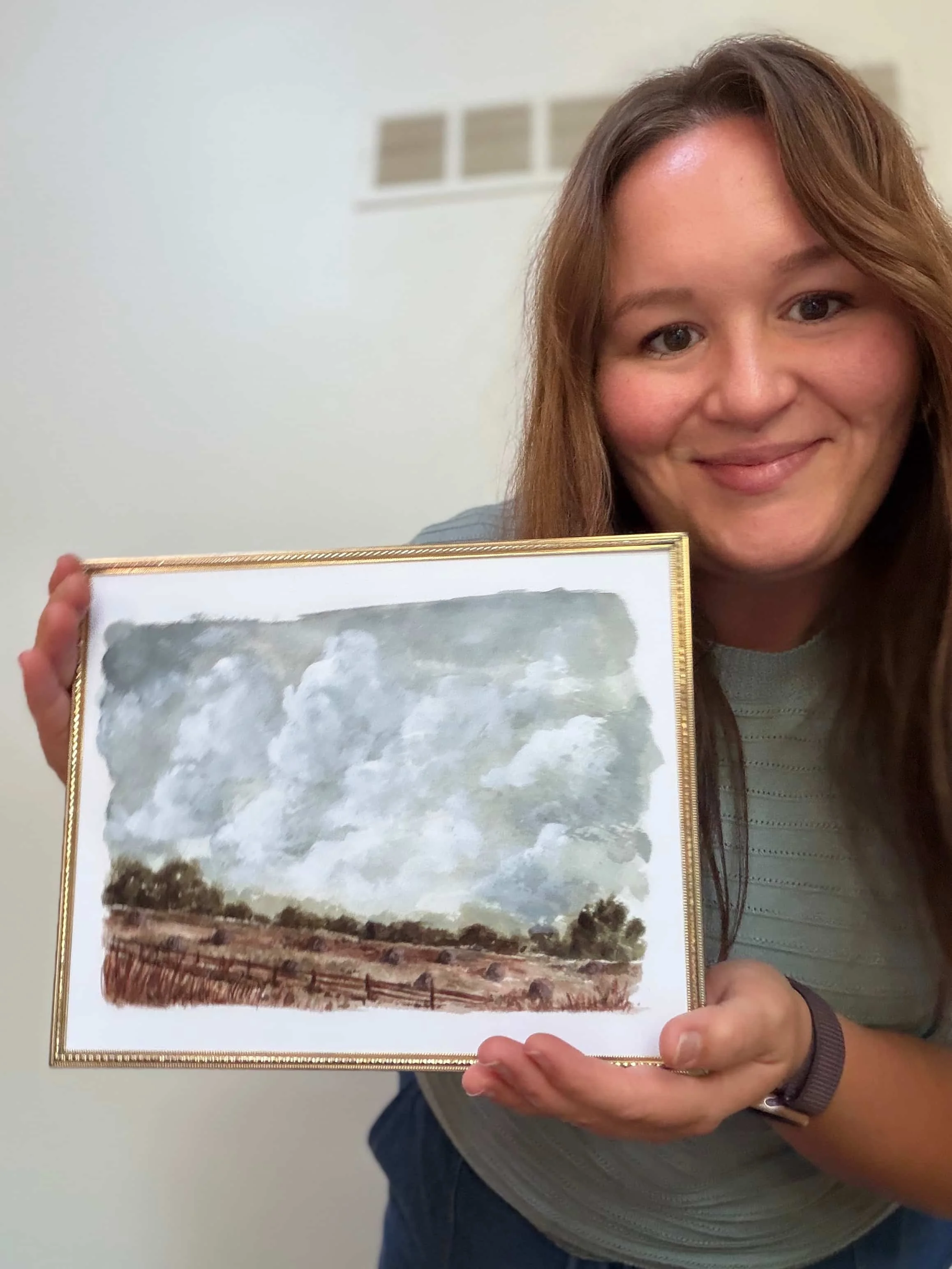 Jess, Indiana watercolor artist and owner of Art and Home co., smiling and holding a framed watercolor landscape painting