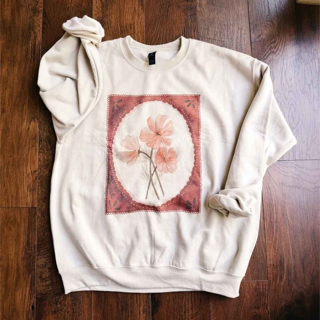 Fall floral watercolor sweatshirt: sand crewneck with pink and peach cosmos in a rust scalloped border, laid flat on a wood floor