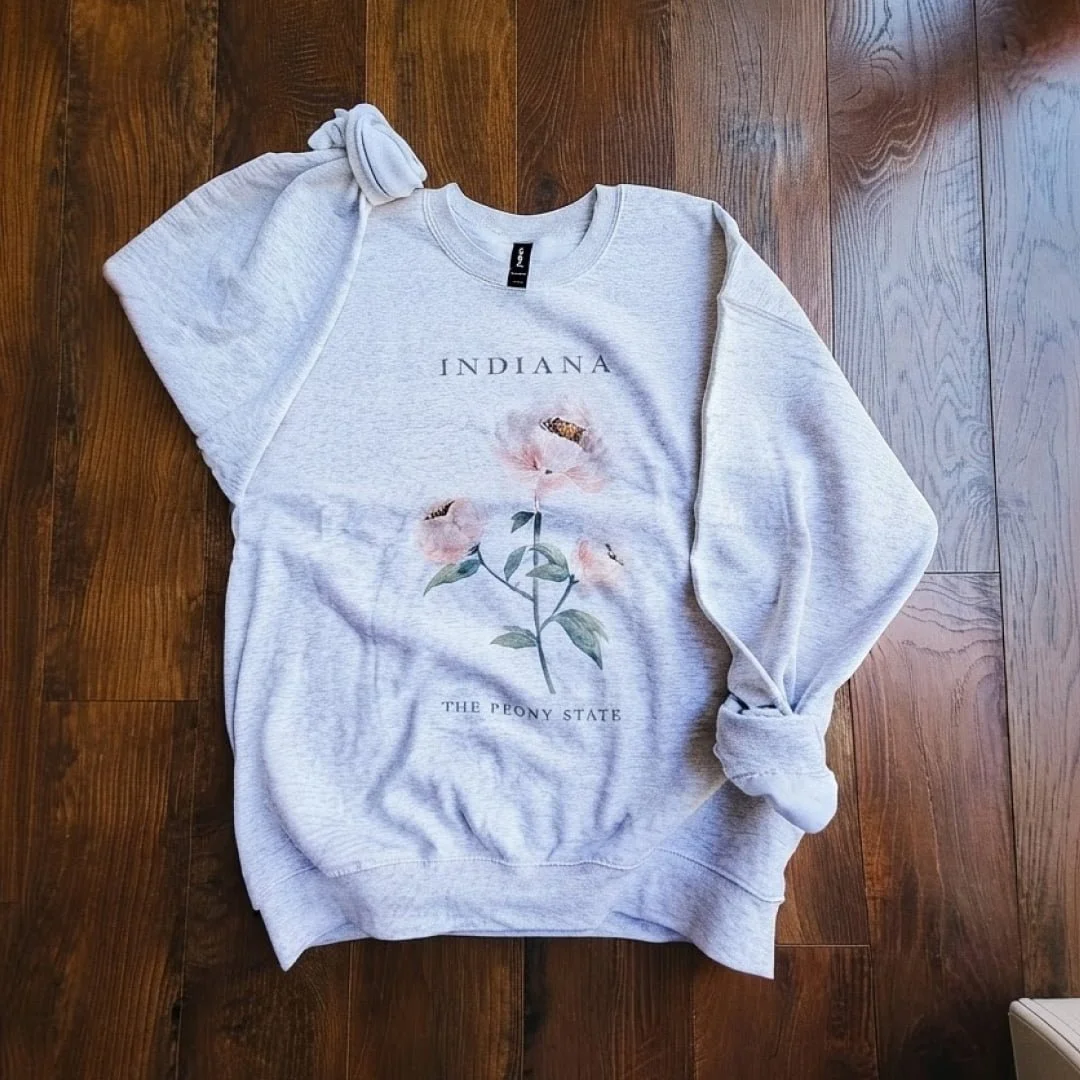 Pink peony watercolor sweatshirt: ash gray crewneck with INDIANA, The Peony State, and a hand-painted pink peony, laid flat on a wood floor