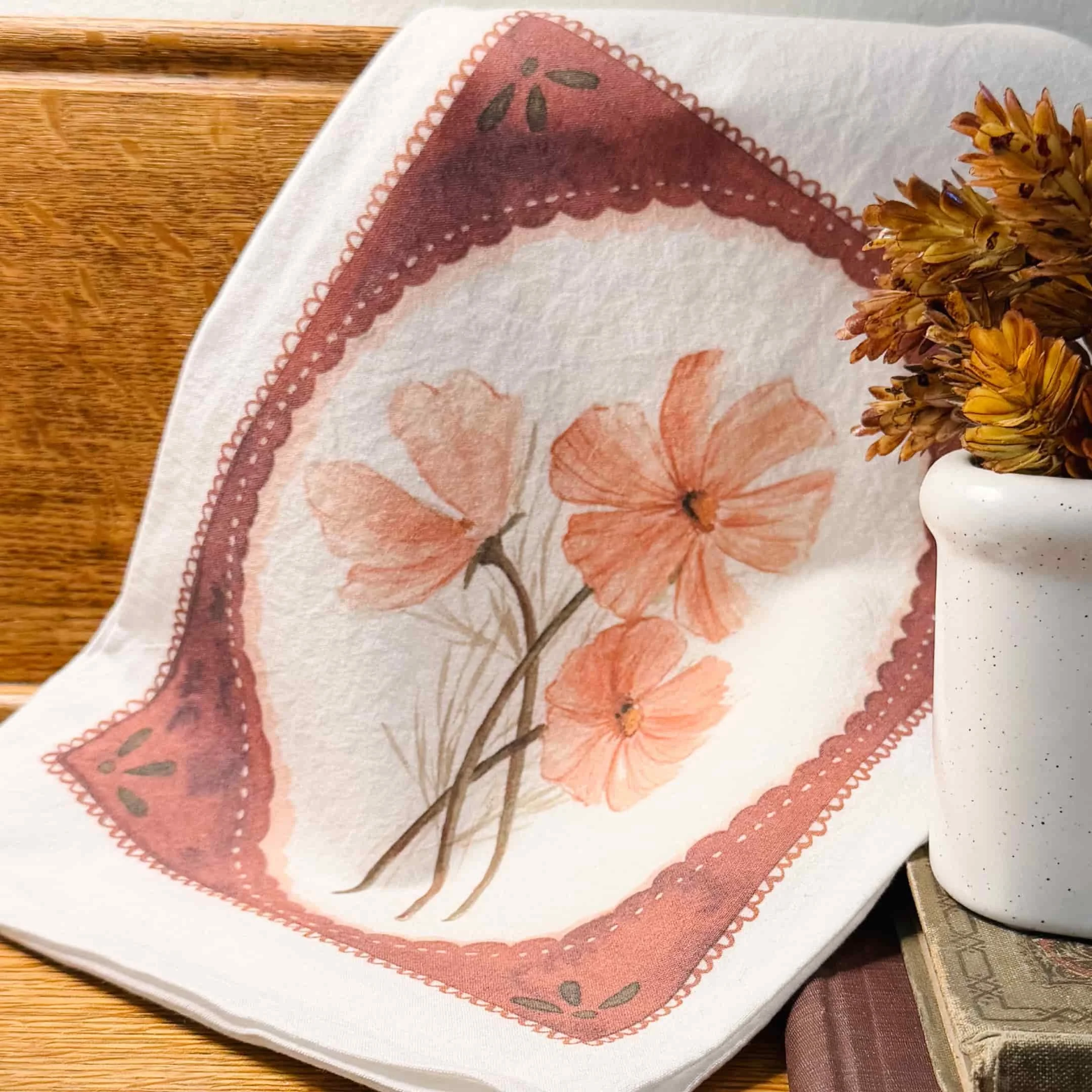 Pink cosmos watercolor tea towel draped on a wooden table beside an antique book and dried fall flowers