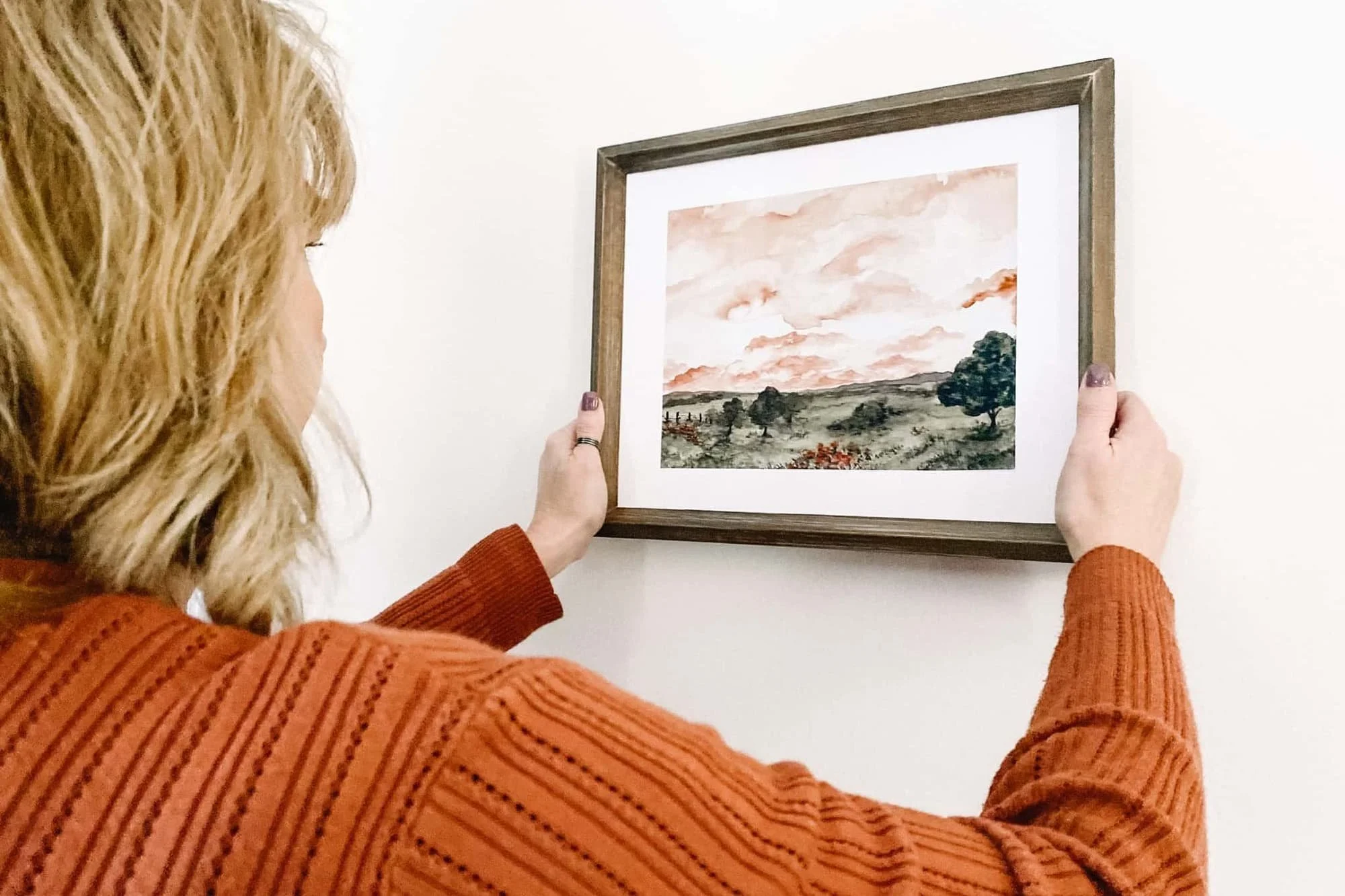Woman in a rust-colored sweater holding a framed watercolor landscape print with a peach sunset sky against a white wall