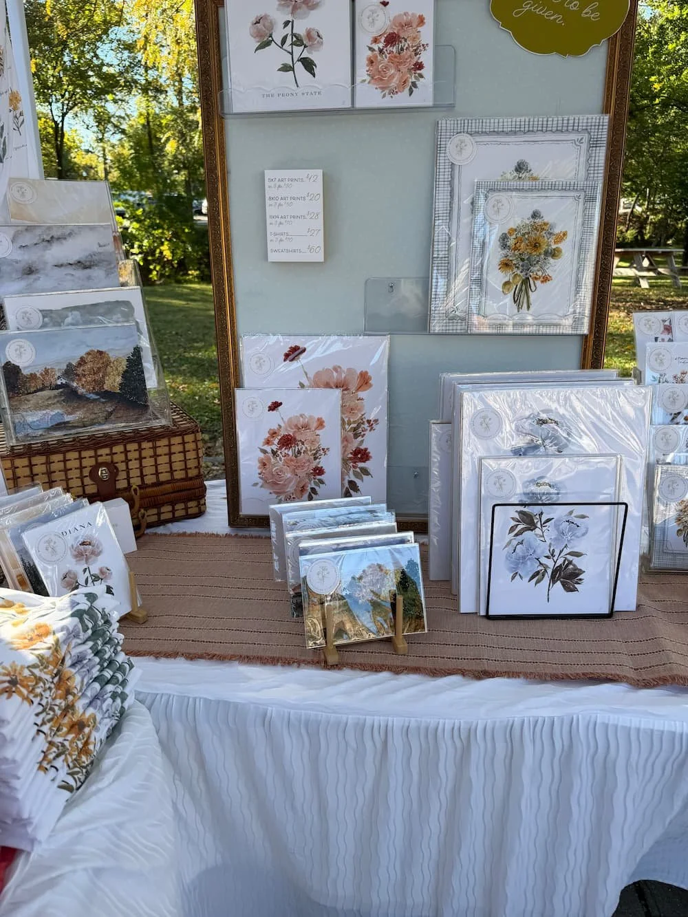 Art and Home co. market booth with hand-painted watercolor art prints, florals, and landscapes displayed on a white-clothed table outdoors