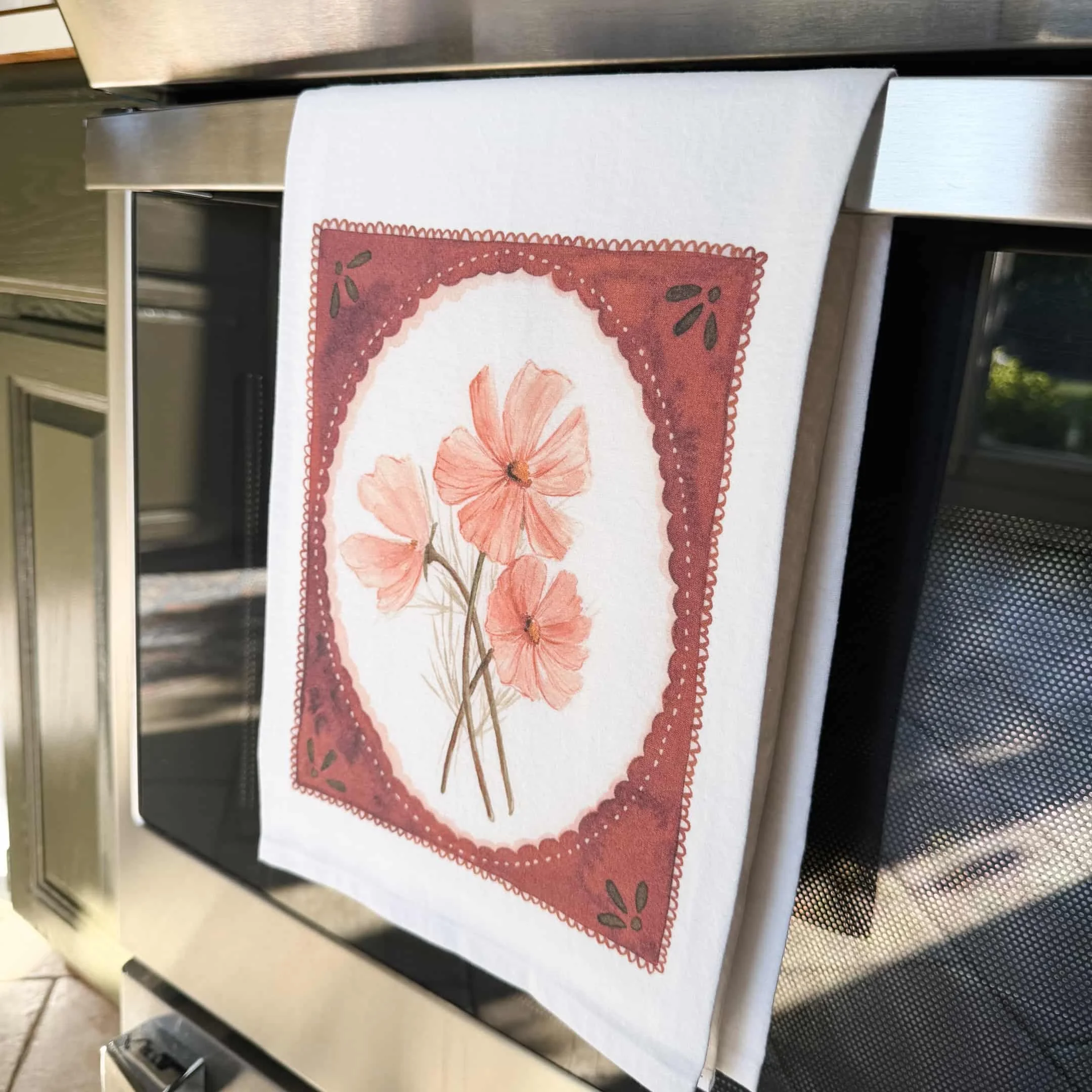 Pink cosmos watercolor tea towel hanging from an oven door handle in a kitchen