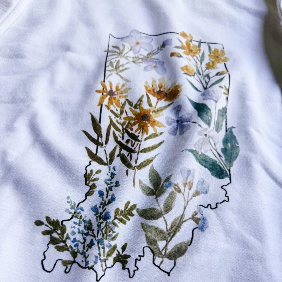 Close-up of watercolor wildflowers inside the Indiana state outline on the white crewneck sweatshirt