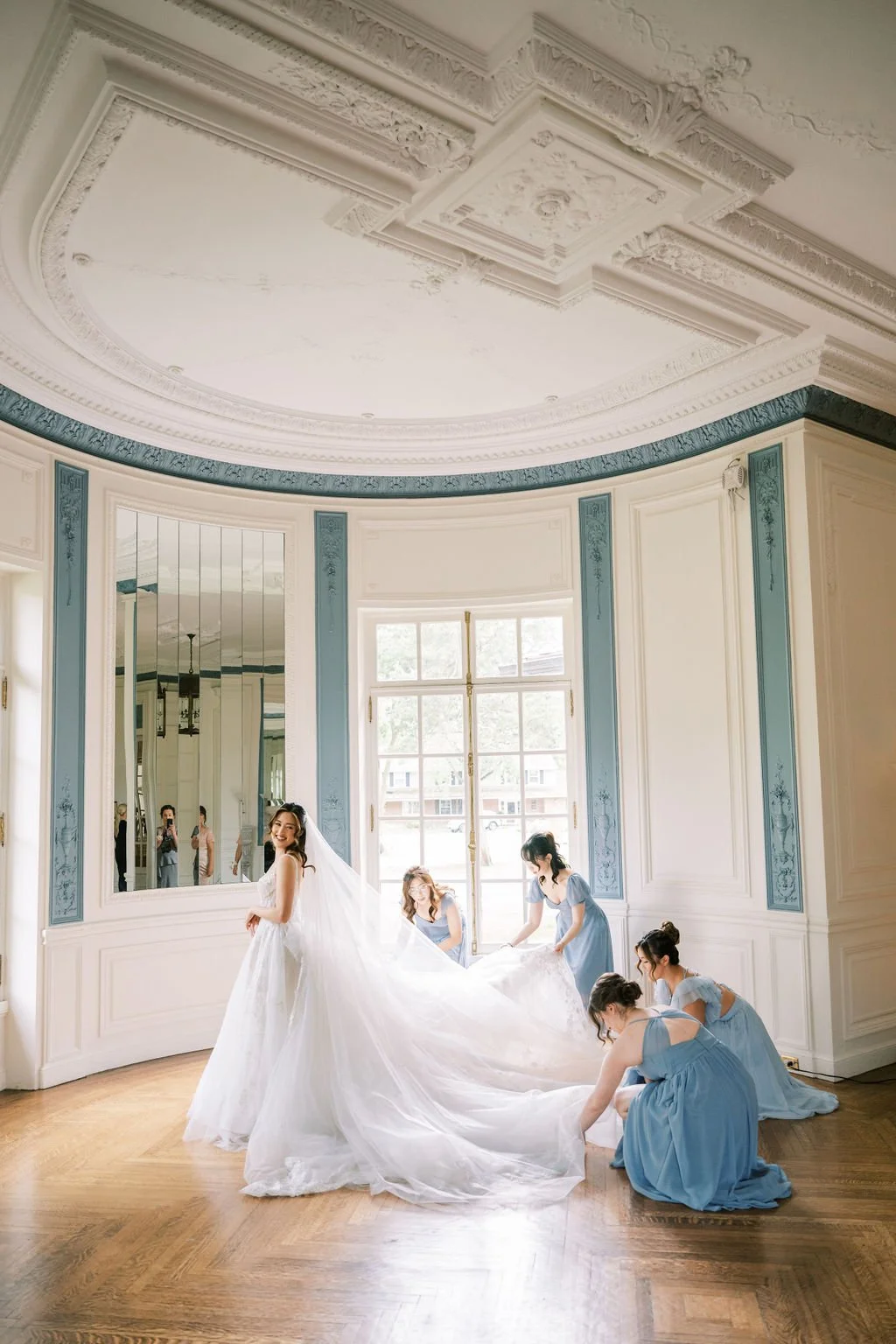 Bride and bridesmaids in dusty blue fixing her veil and gown in the mirror.