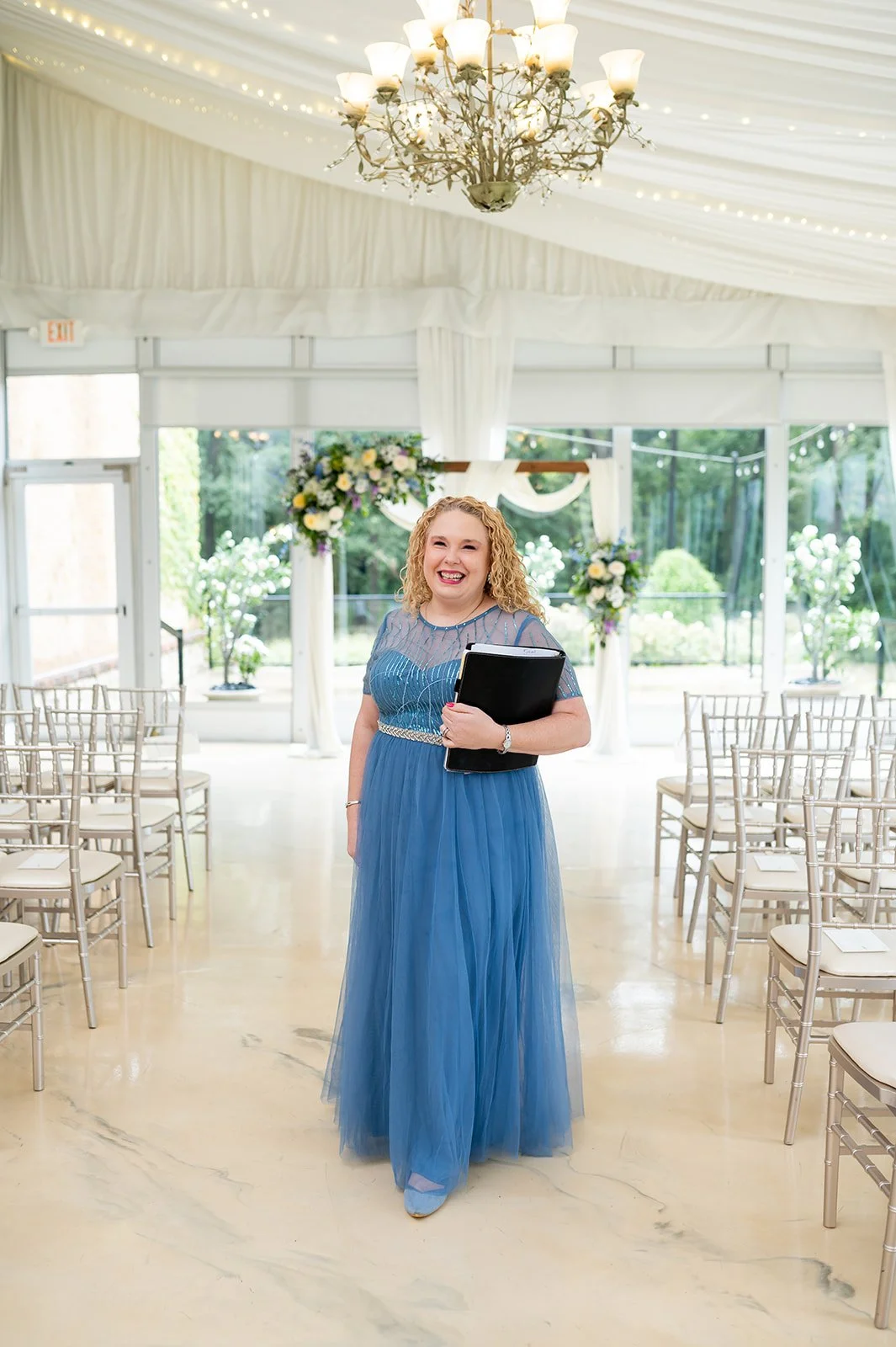 Professional Officiant Carla Doshi of Forever Incredible Ceremonies stands at The Monte Bello Estate in a blue dress with her ceremony binder.