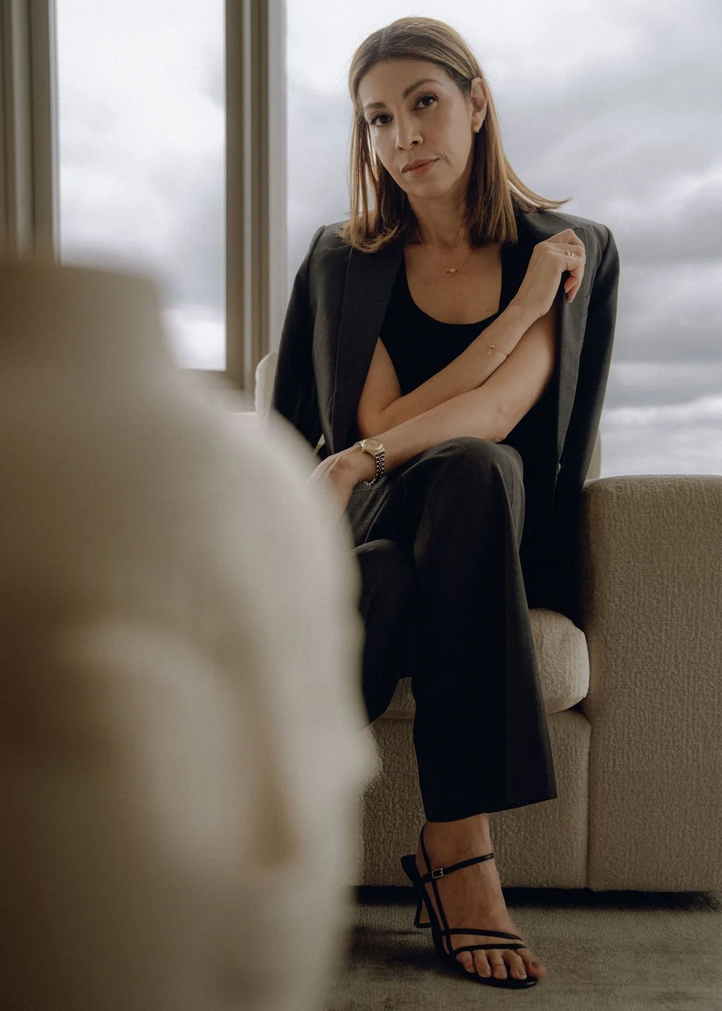 Christina DiStefano sitting on a beige sofa near a window with cloudy sky, wearing a black dress, blazer, high heels, gold jewelry, and a watch, looking at the camera.