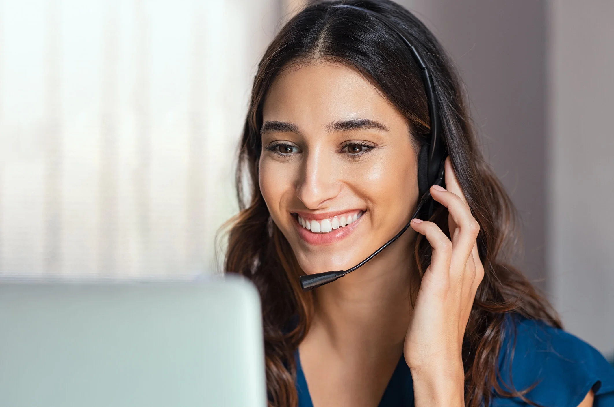 A young woman with long brown hair wearing a headset and smiling while looking at a computer screen.