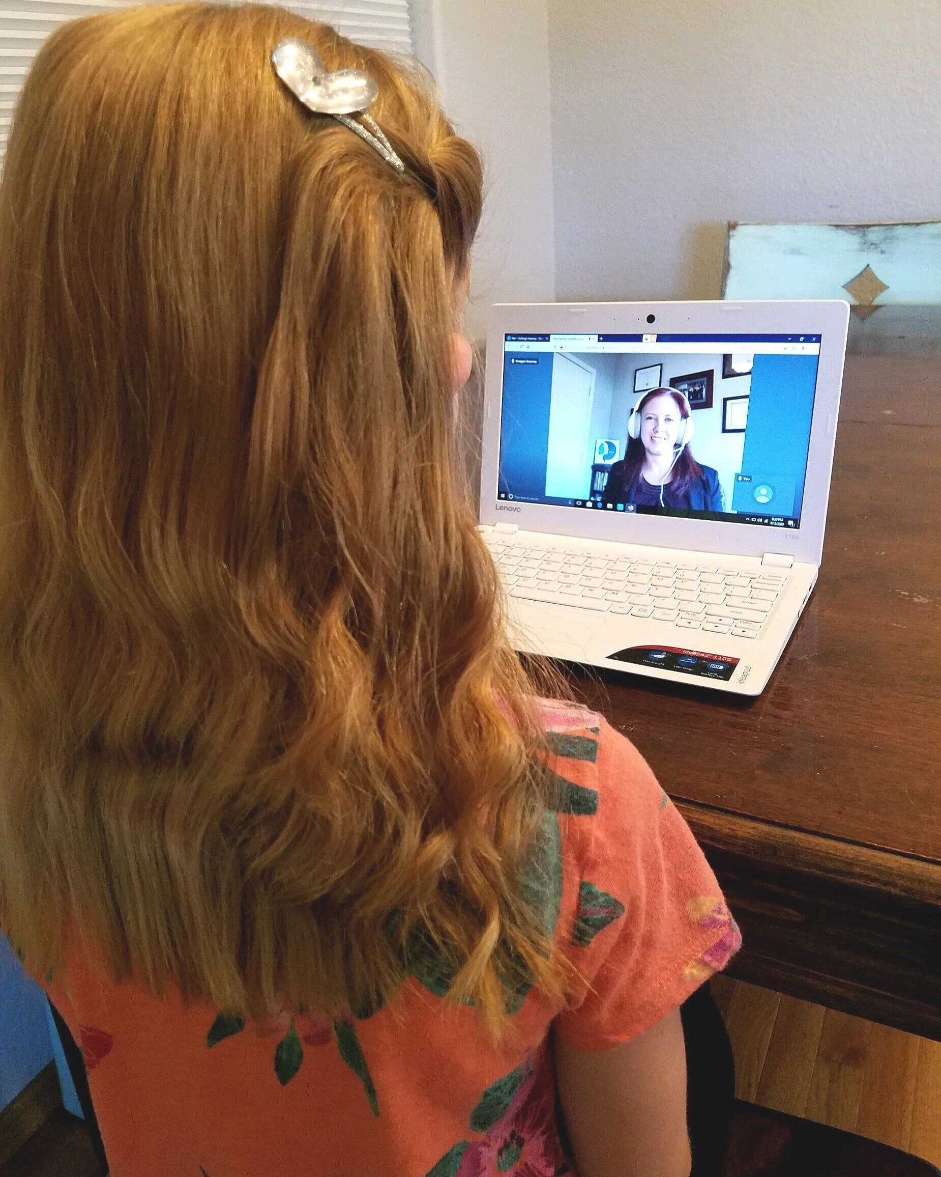 A girl with long, curly red hair wearing a pink shirt with a floral pattern, sitting at a wooden table, participating in a video call on a white Lenovo laptop with another woman on the screen, who has headphones and is smiling.