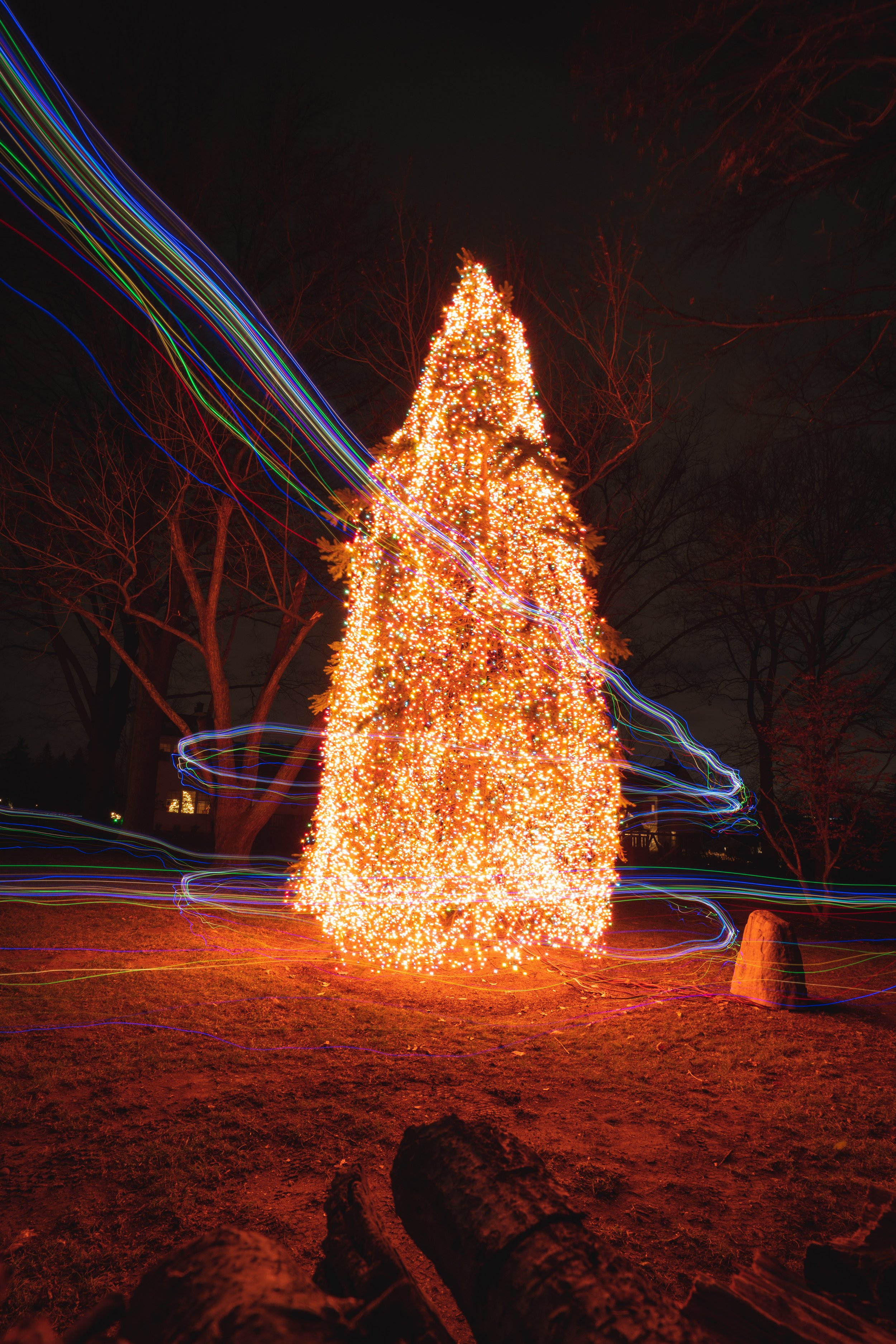A Christmas tree decorated with numerous bright orange and yellow lights, with colorful light trails swirling around it at night outdoors.