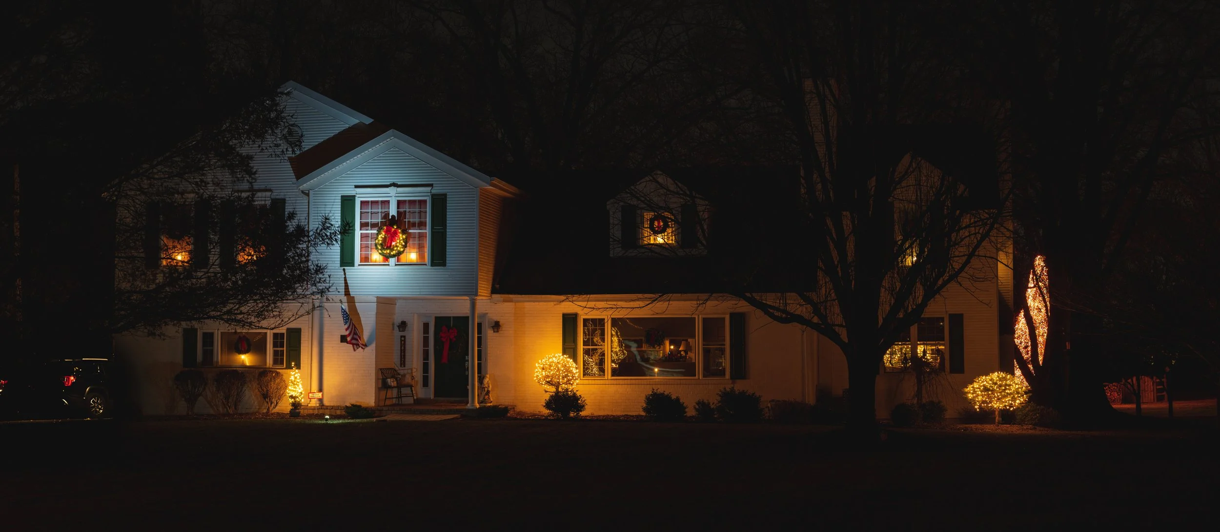 A house decorated with Christmas lights and wreaths at night, with trees and bushes illuminated around it.