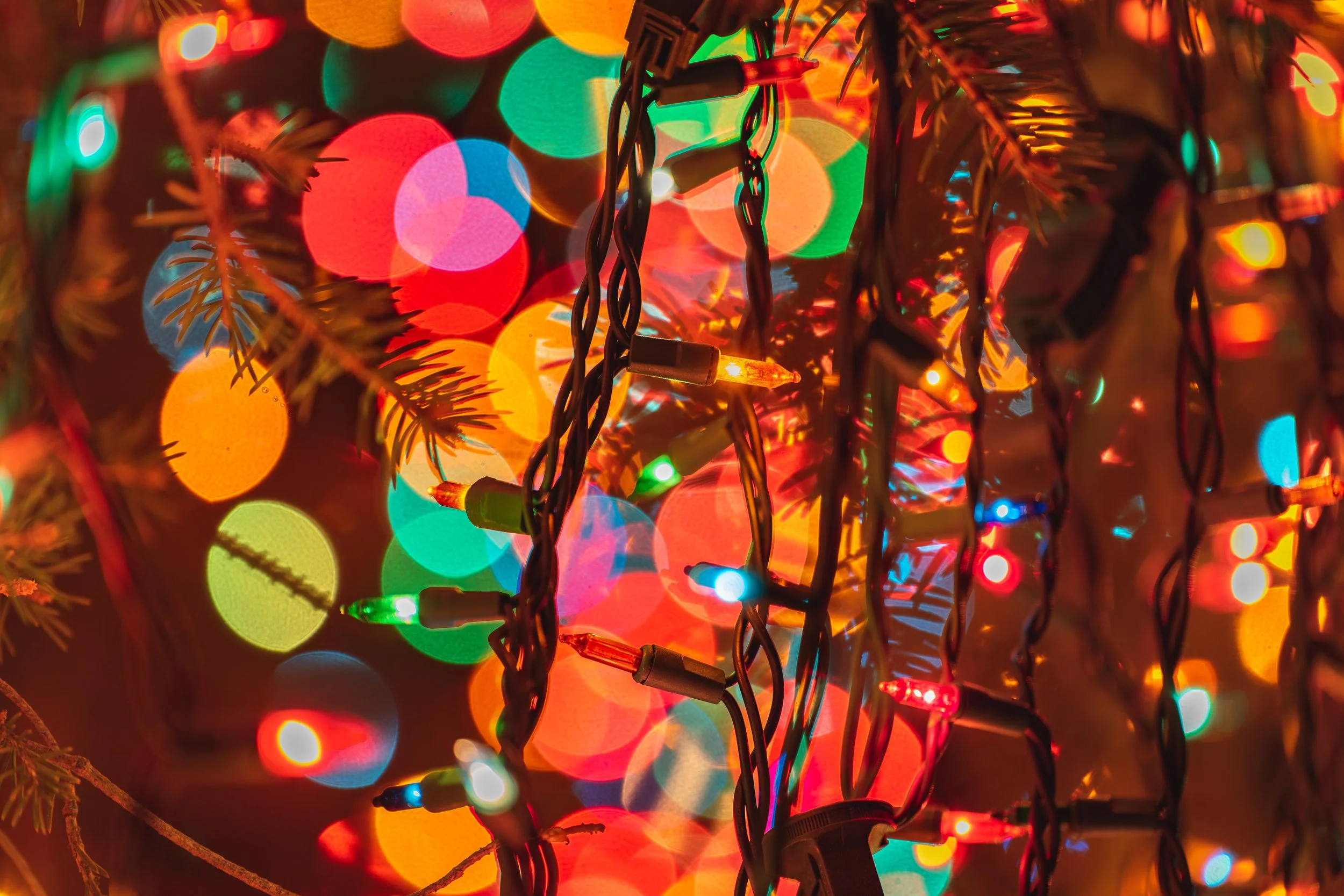 Close-up of multicolored Christmas string lights hanging in front of a blurred background of multicolored bokeh lights.