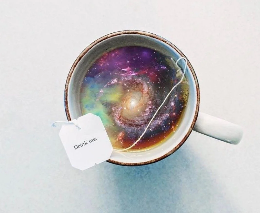 A cup filled with coffee that has a galaxy and nebula pattern, creating the illusion of a galaxy inside the coffee. There is a tea bag with a tag labeled 'Drink me' and a string placed across the cup.