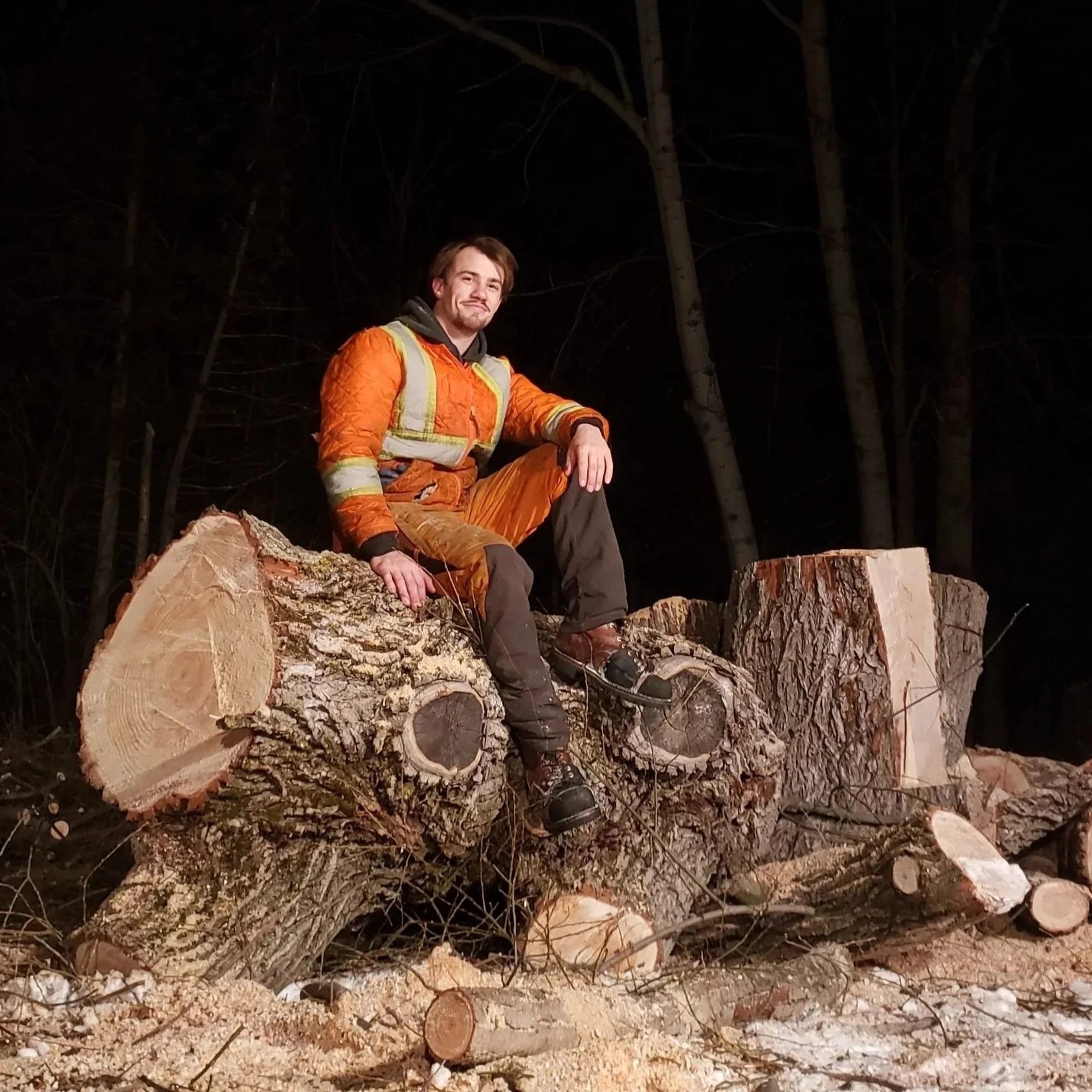 A man dressed in orange outdoor gear sits on a large felled tree trunk in a nighttime forest setting, with dark trees in the background.