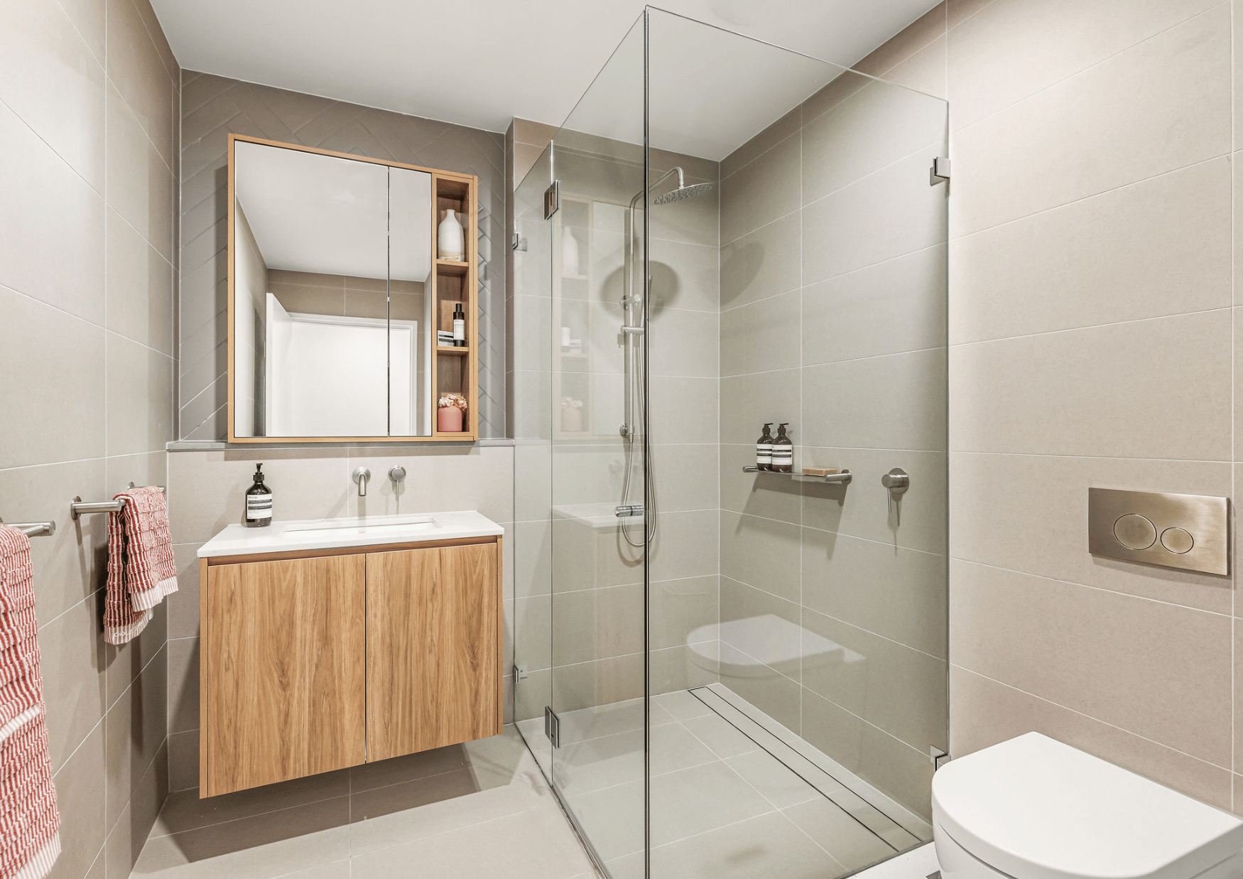 Modern bathroom with beige tiles, a wooden vanity with a sink, a mirror cabinet, and a glass-enclosed shower area with a rain shower head. There are red and white towels hanging on a towel bar and two black soap dispensers on the vanity and shower sh