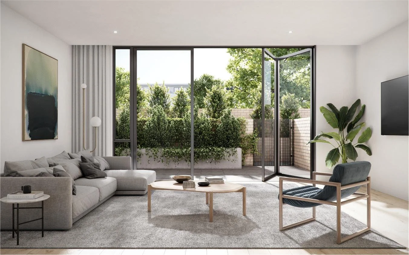 A modern living room with large glass sliding doors opening to a green outdoor patio. The room has a light gray sectional sofa, a wooden armchair with dark cushions, a round coffee table, and a small side table. There is an abstract painting on the w