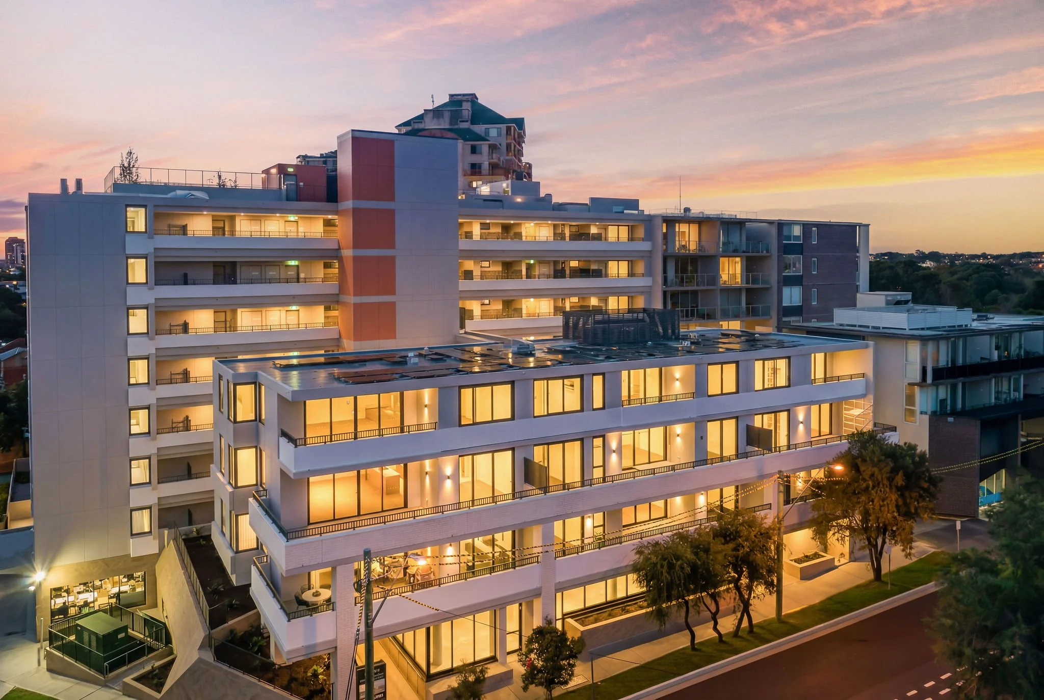 Tall modern apartment building at sunset with lit windows, balconies, rooftop garden, and trees in the foreground.